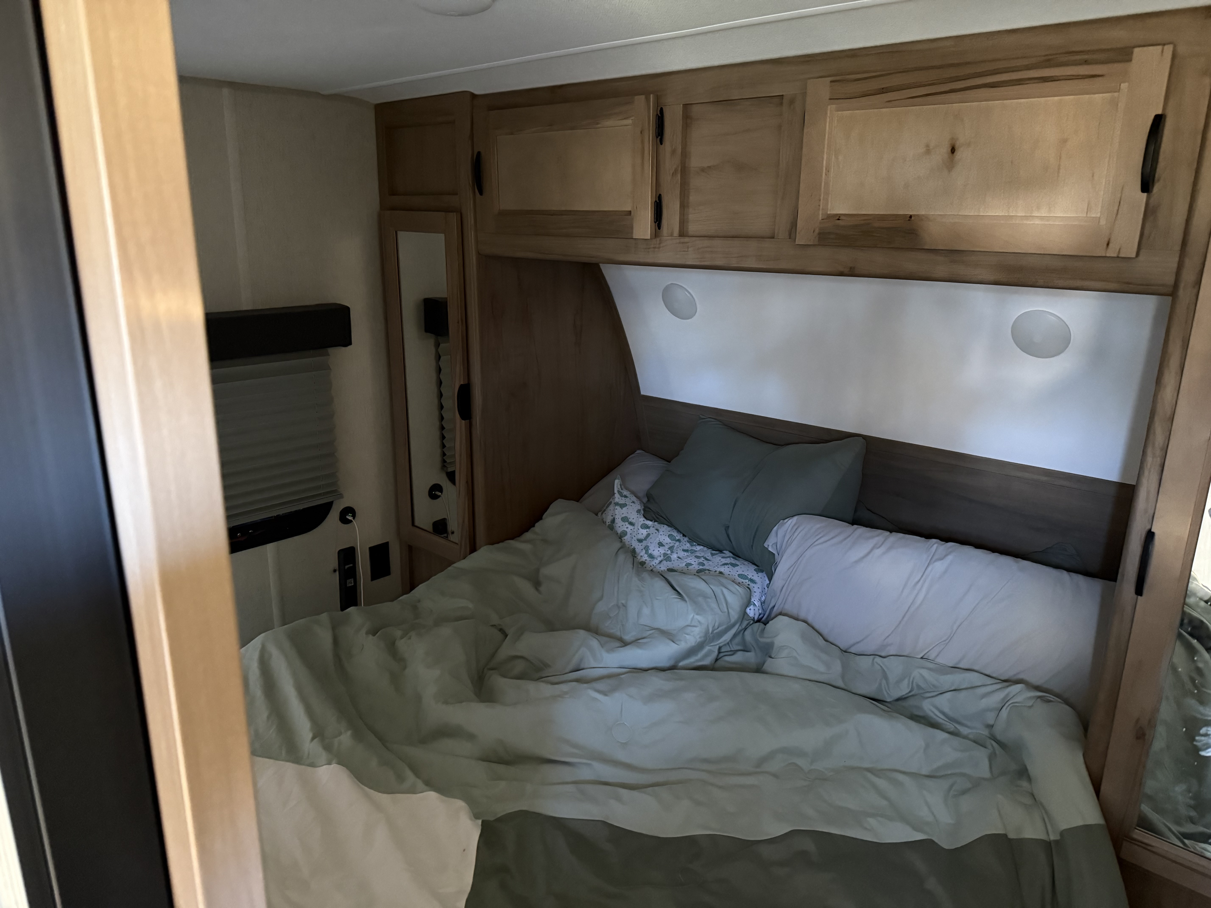 Cozy RV rental bedroom featuring a bed with light green and white bedding, overhead wooden cabinets, and a bedside mirror.. Forest River Cherokee Grey Wolf 2024