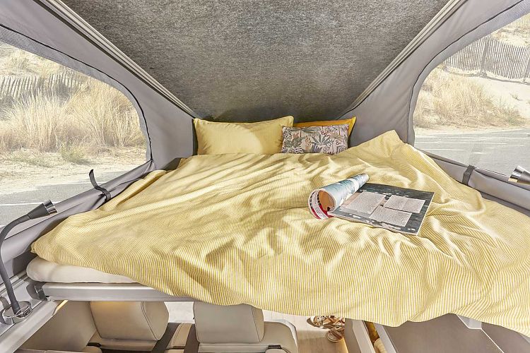 Comfortable pop-top bed in an RV rental with yellow striped bedding, offering views of a road and grassy dunes.. Westfalia Kelsey 2024