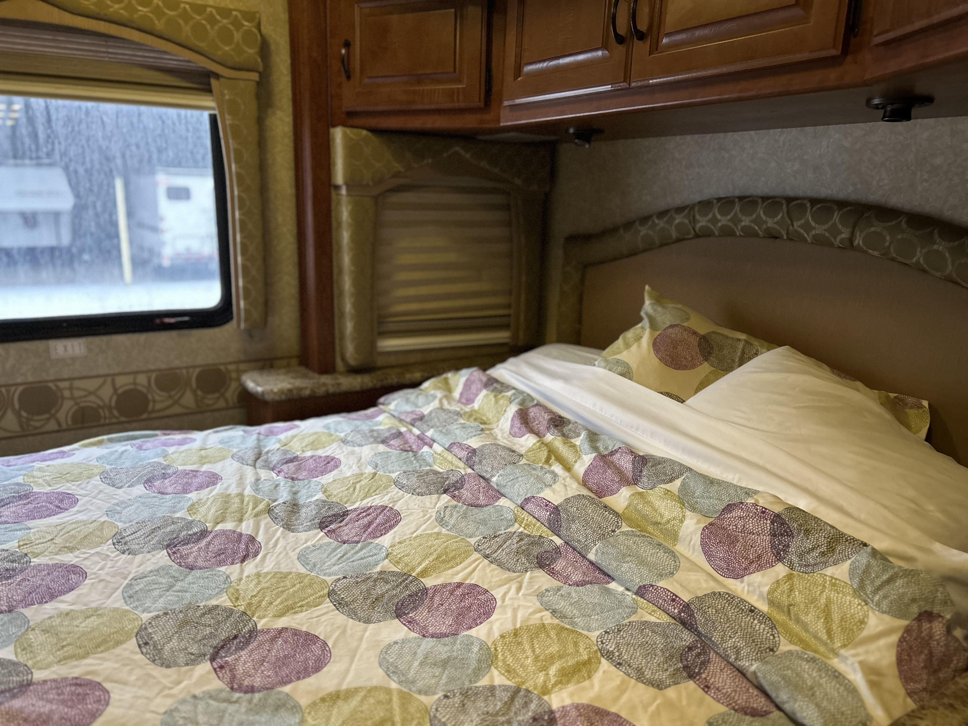 Cozy rv rental bedroom with a colorful comforter, pillows, and a window showing trailers outside in snowy weather.. Four Winds Hurricane 2010