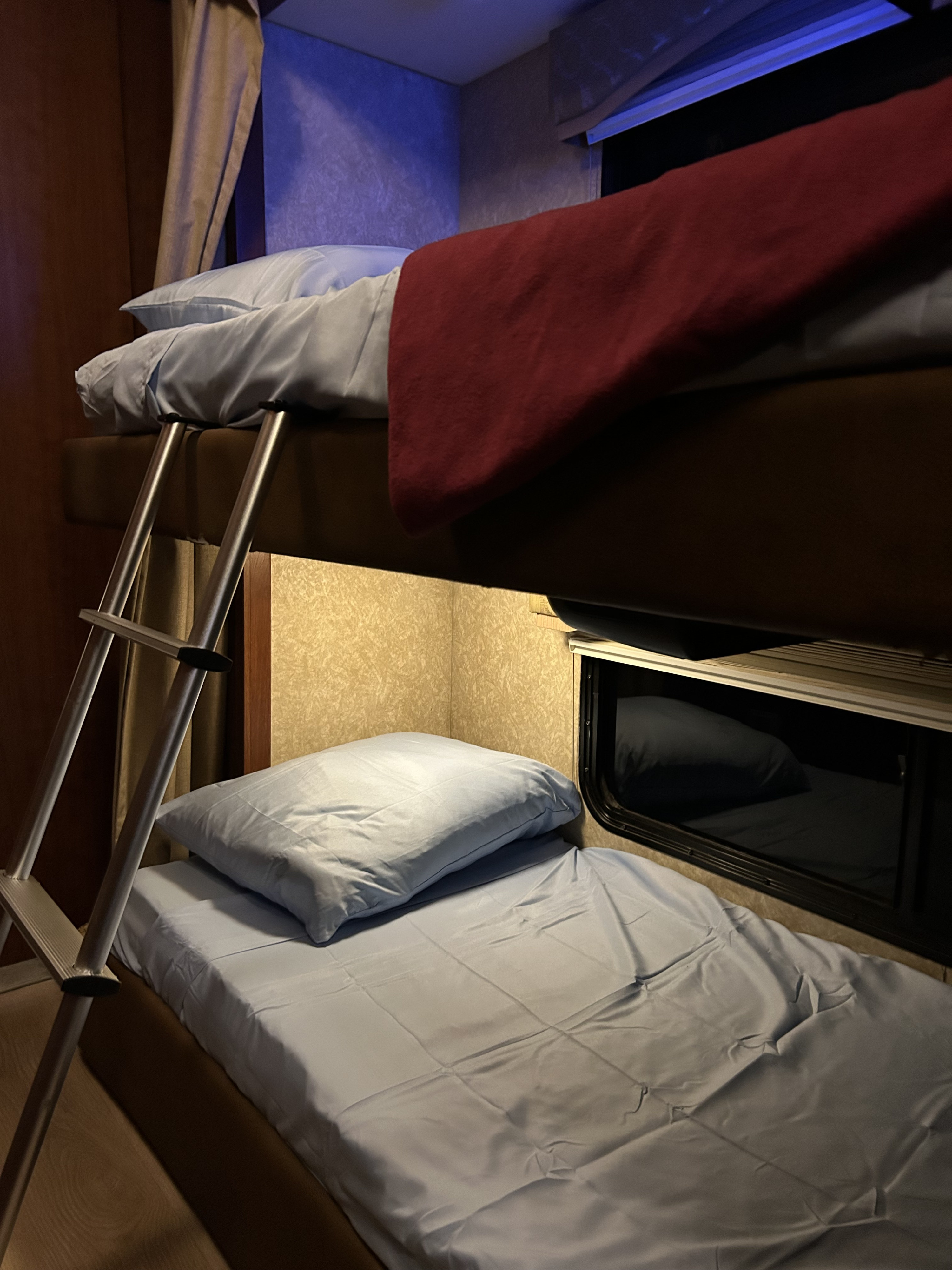 Compact RV rental bunk beds with blue sheets and pillows, a red blanket on the top bunk, and a metal ladder.. Four Winds Hurricane 2010
