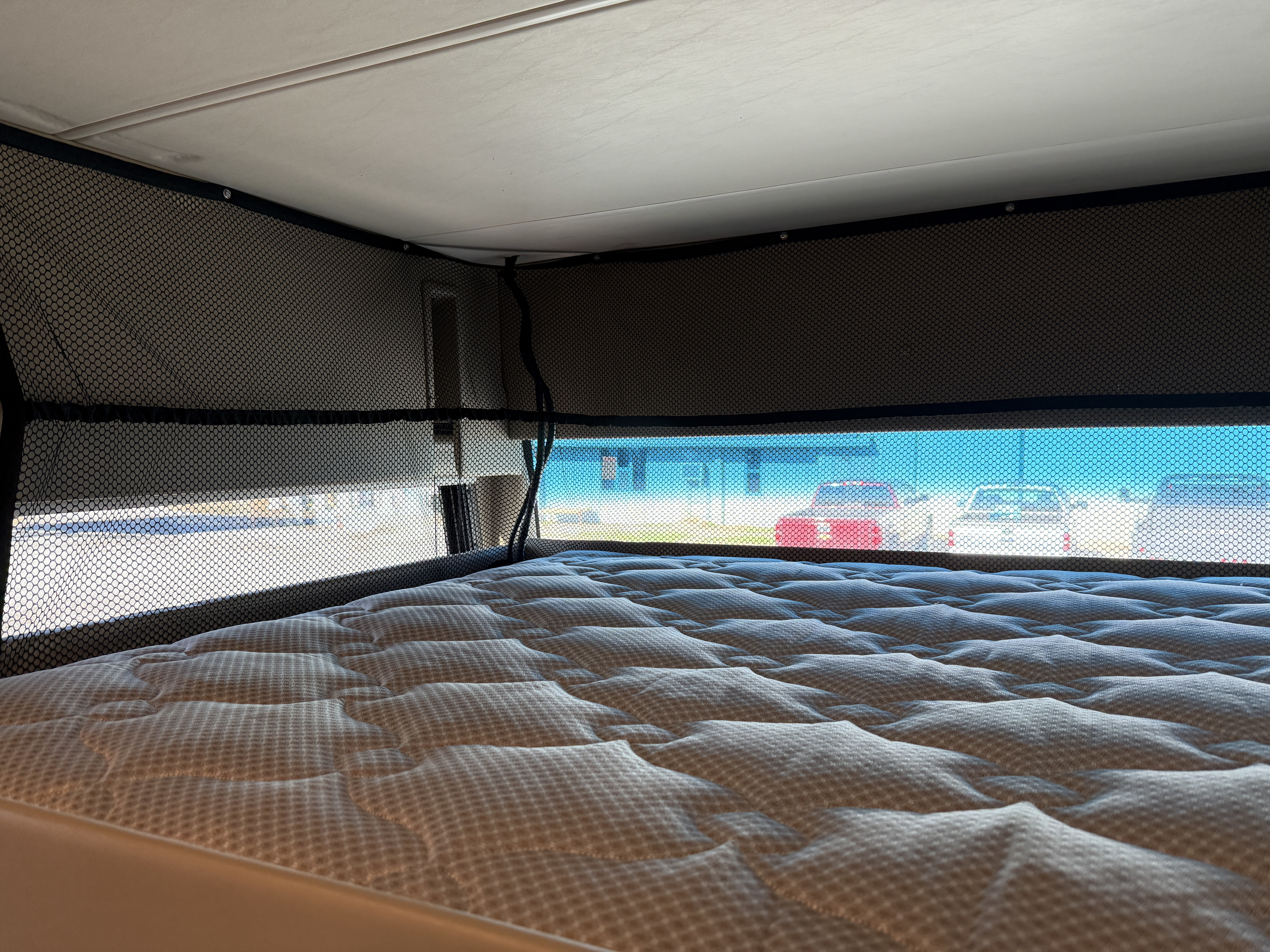 RV rental bed area: quilted mattress and mesh windows with a view of a parking lot outside.. Ford Thor Challenger 2021