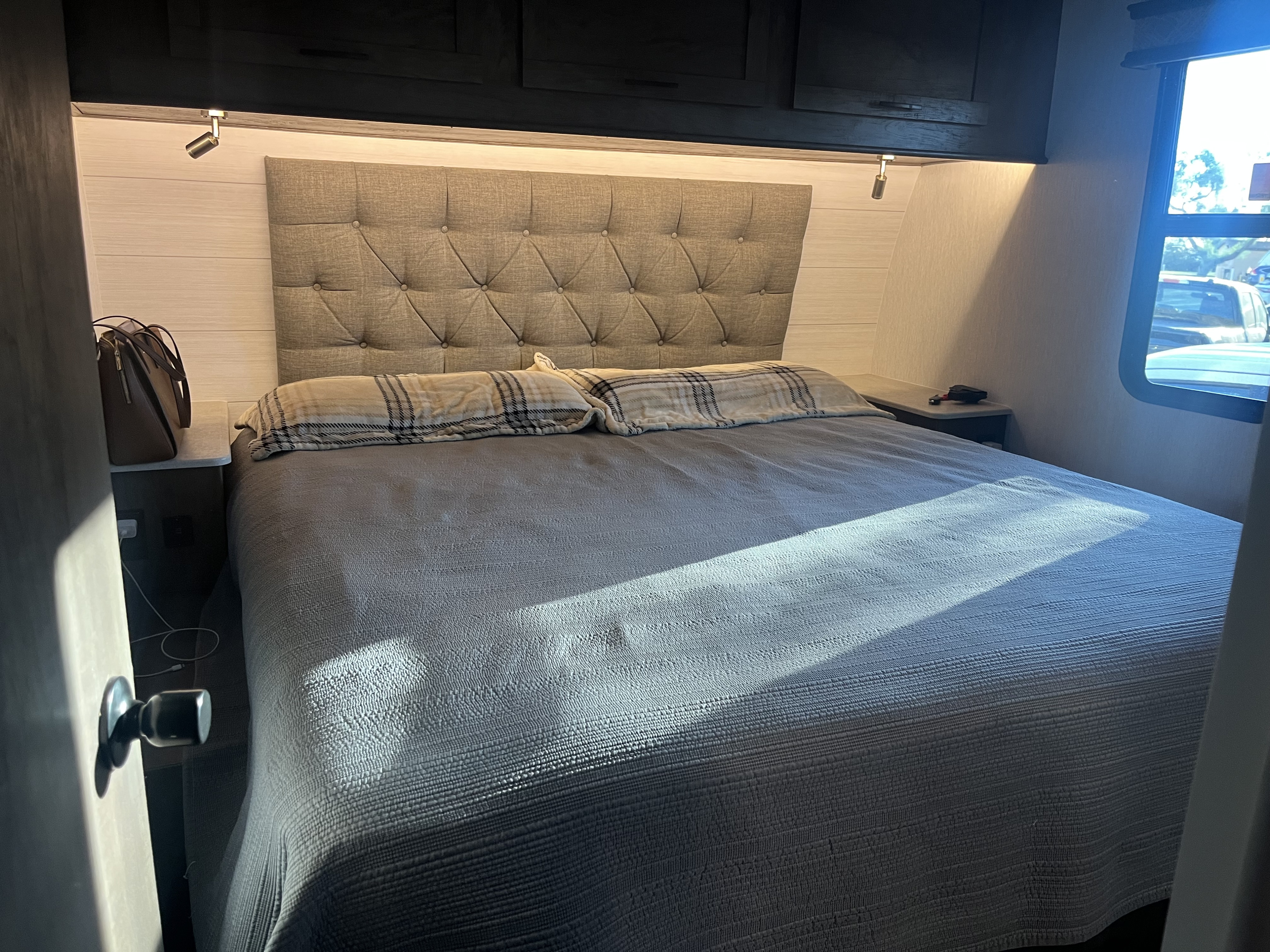 An RV rental bedroom features a comfy bed with a tufted headboard, two plaid pillows, and natural light streaming from a window.. Forest River Sandstorm 2022