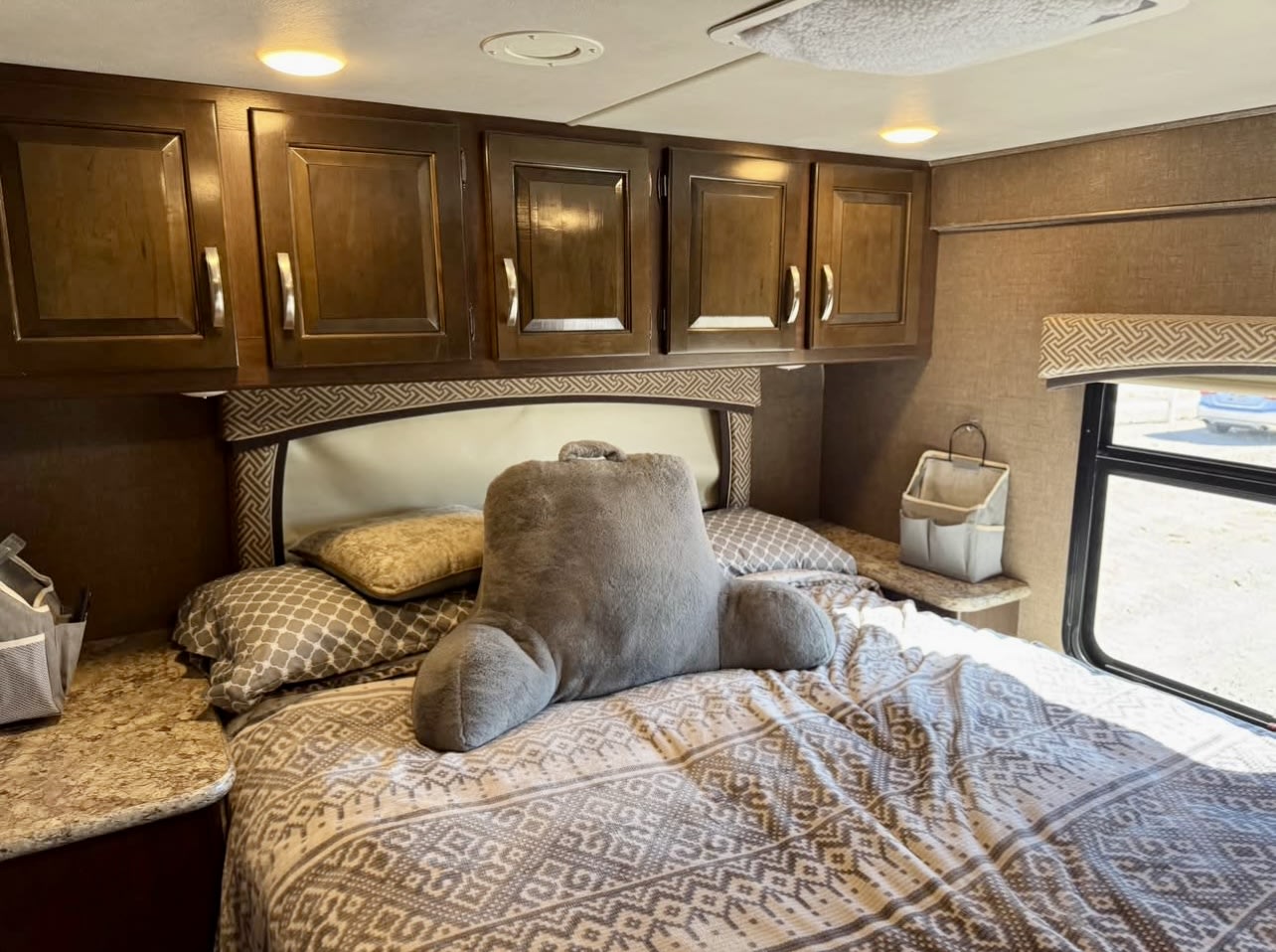Inside an RV rental bedroom: cozy bed with patterned sheets, overhead storage, and a reading pillow. Comfortable travels await.. Thor Chateau 2017