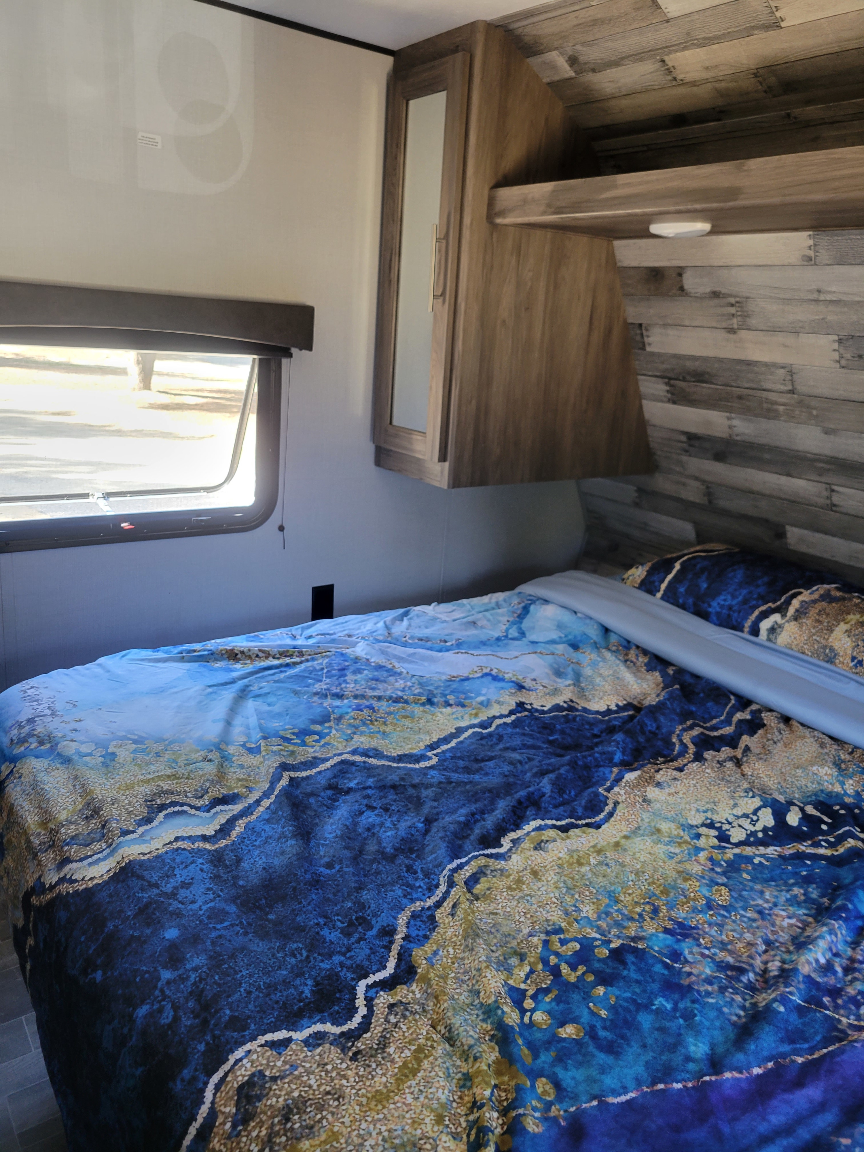 Inside an RV rental, this cozy bedroom features a bed with vibrant blue & gold bedding, a window, and wood cabinetry.. Crossroads RV Zinger 2021