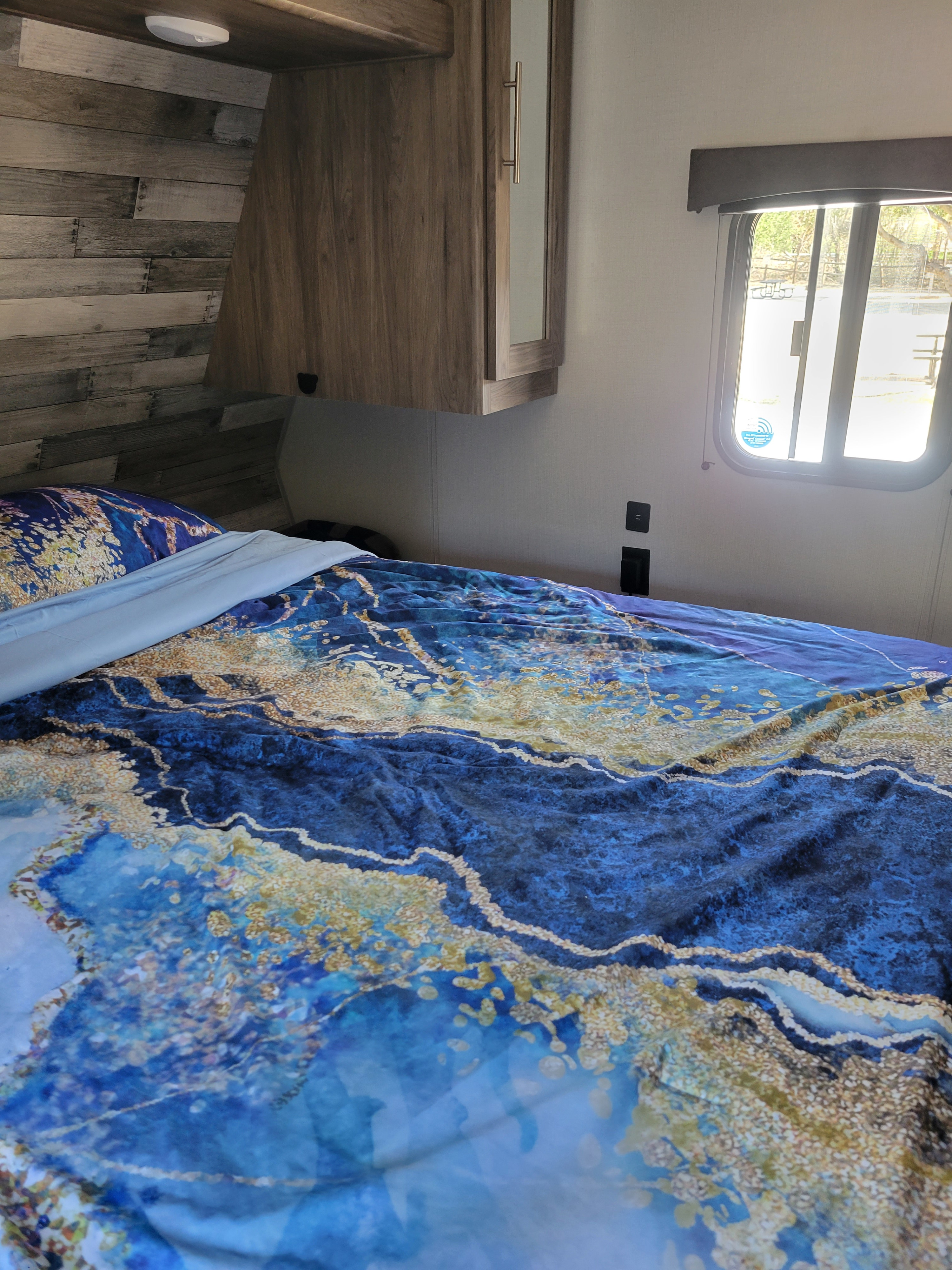 Comfortable RV rental bedroom with blue and gold patterned bedding, wooden headboard, and a sunlit window.. Crossroads RV Zinger 2021