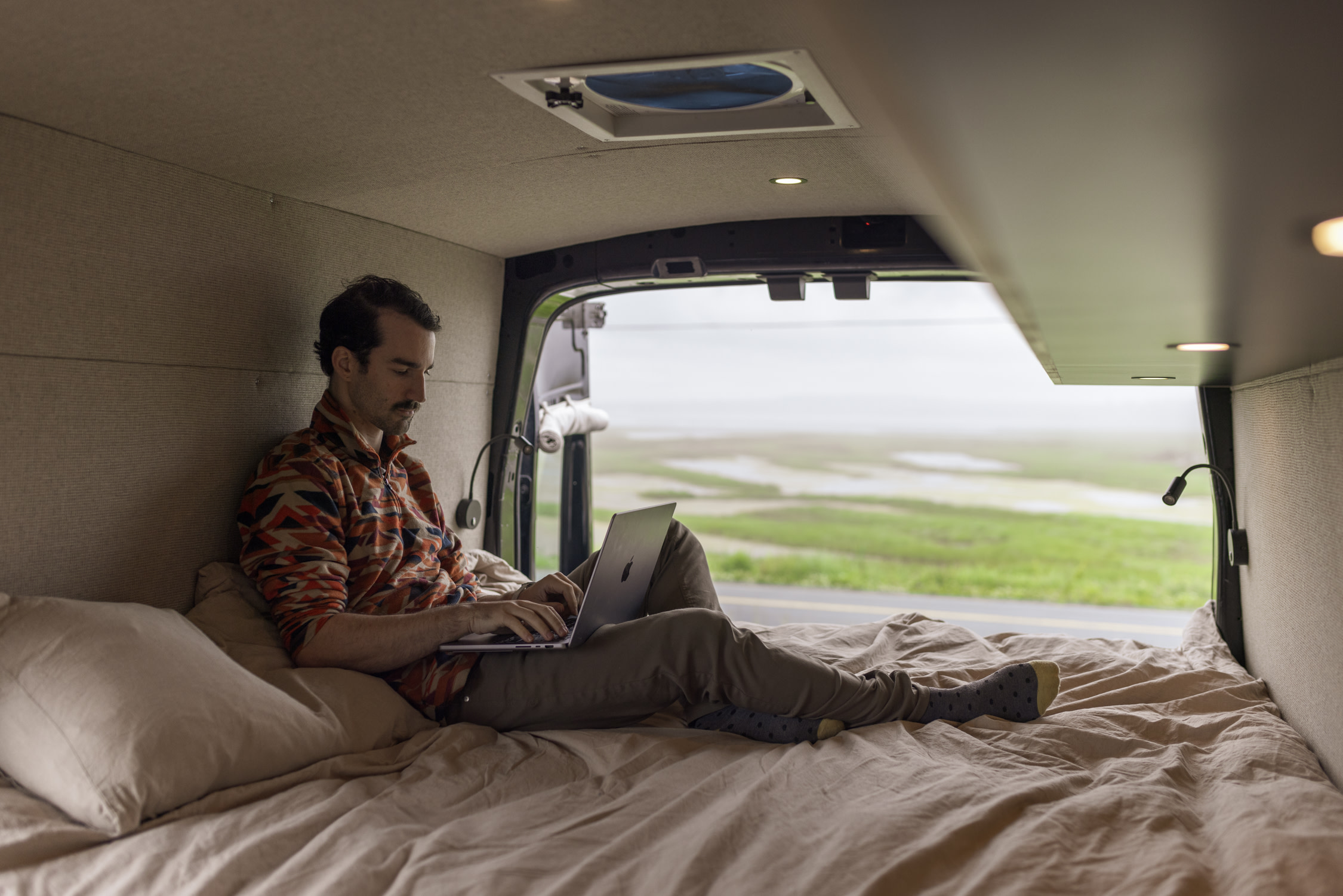 Experience digital nomad life! An RV rental provides a cozy workspace for a man on a bed, enjoying a scenic view.. Ford Transit 2023