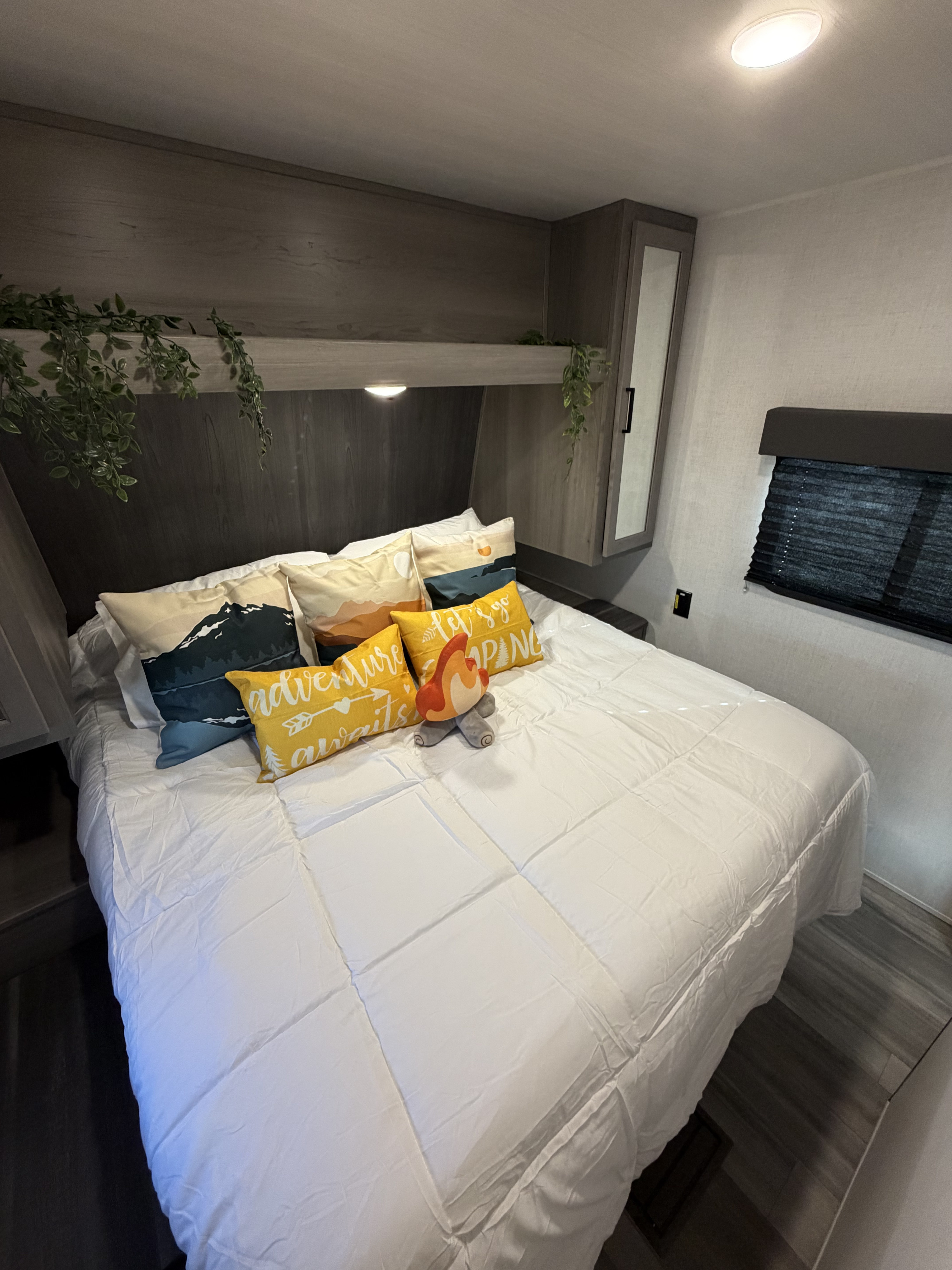 Inside this inviting RV rental, a comfy bedroom features a white bed, camping pillows, and overhead storage with greenery.. Keystone RV Hideout 2024