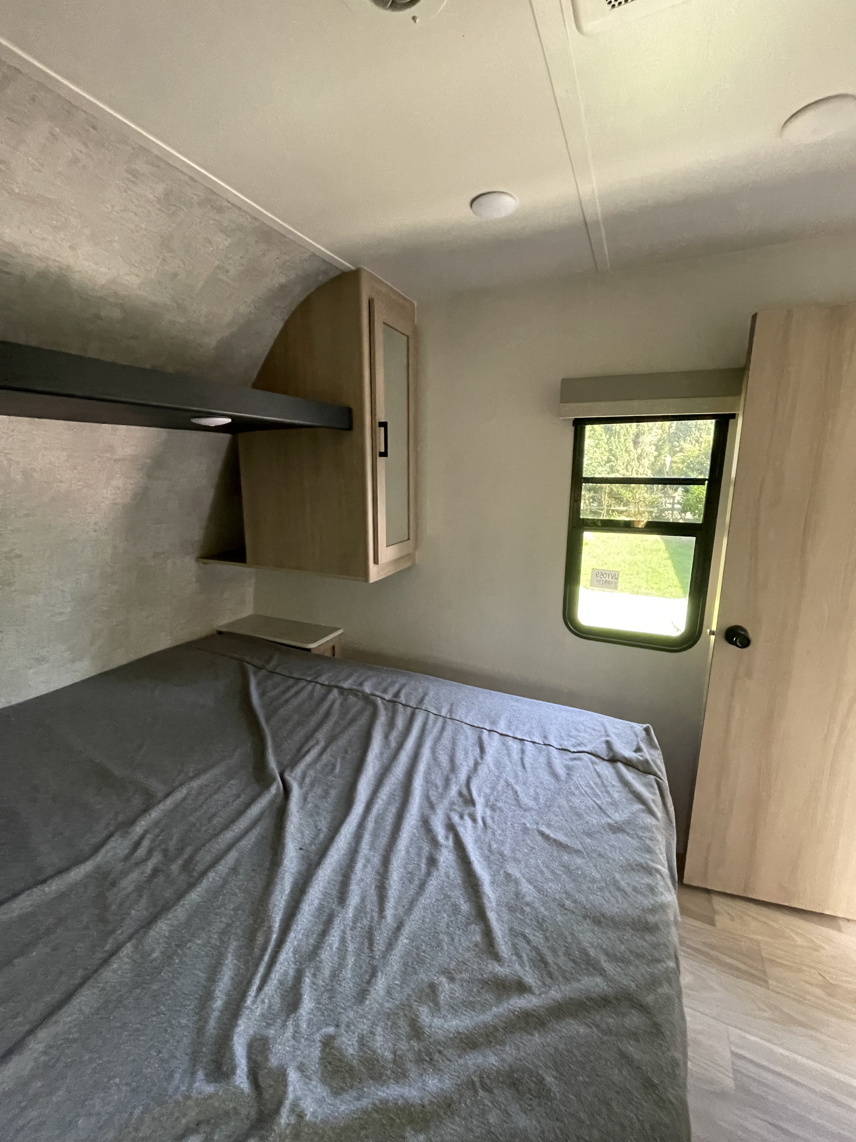 Inside an RV rental, this bedroom features a bed with grey sheets, overhead storage, and a window showcasing a green outdoor view.. Winnebago Voyage 2023
