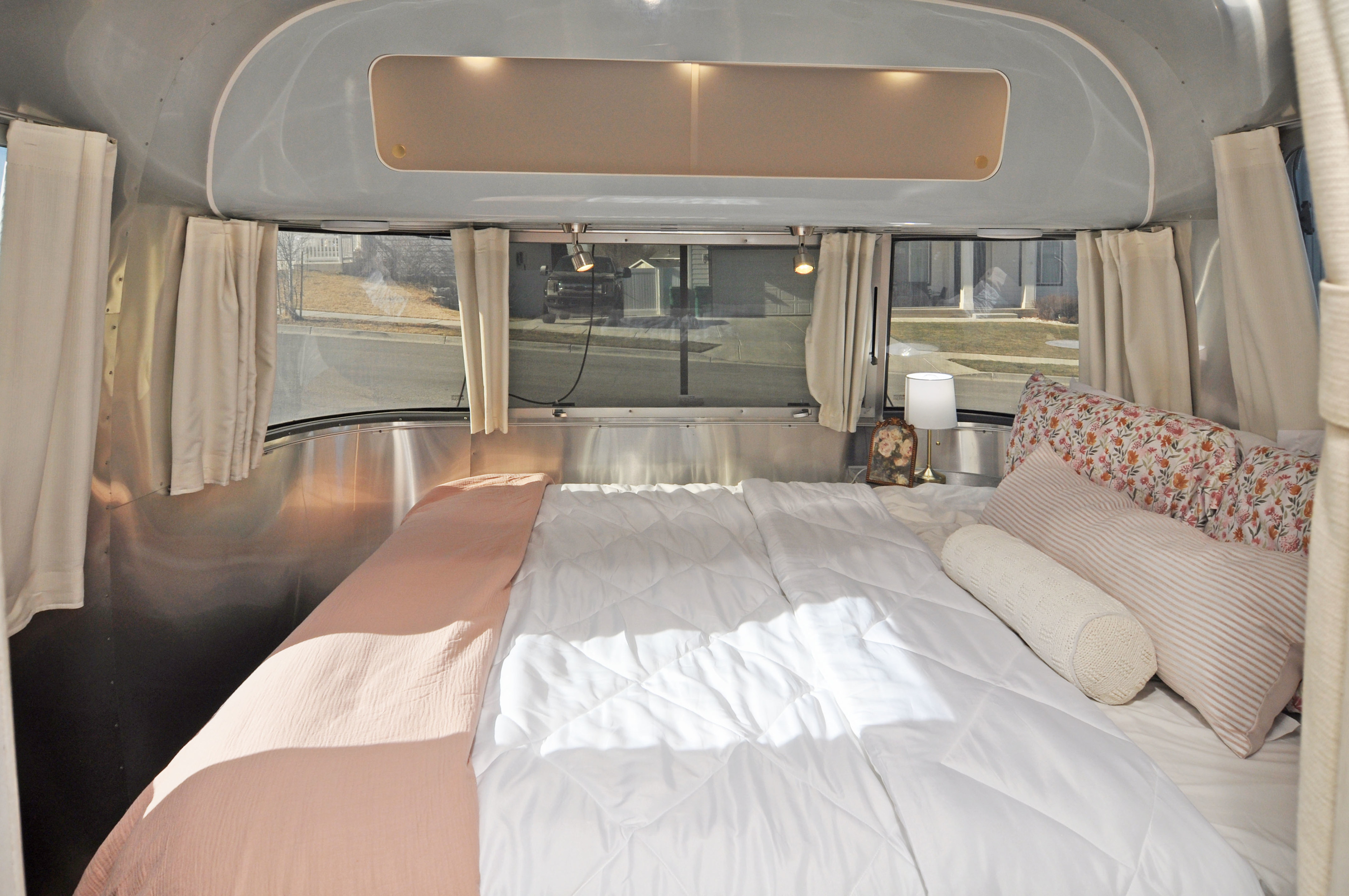 Plush bed in a stylish Airstream RV rental, featuring white & pink bedding, comfy pillows, and panoramic window views.. Airstream International 2021