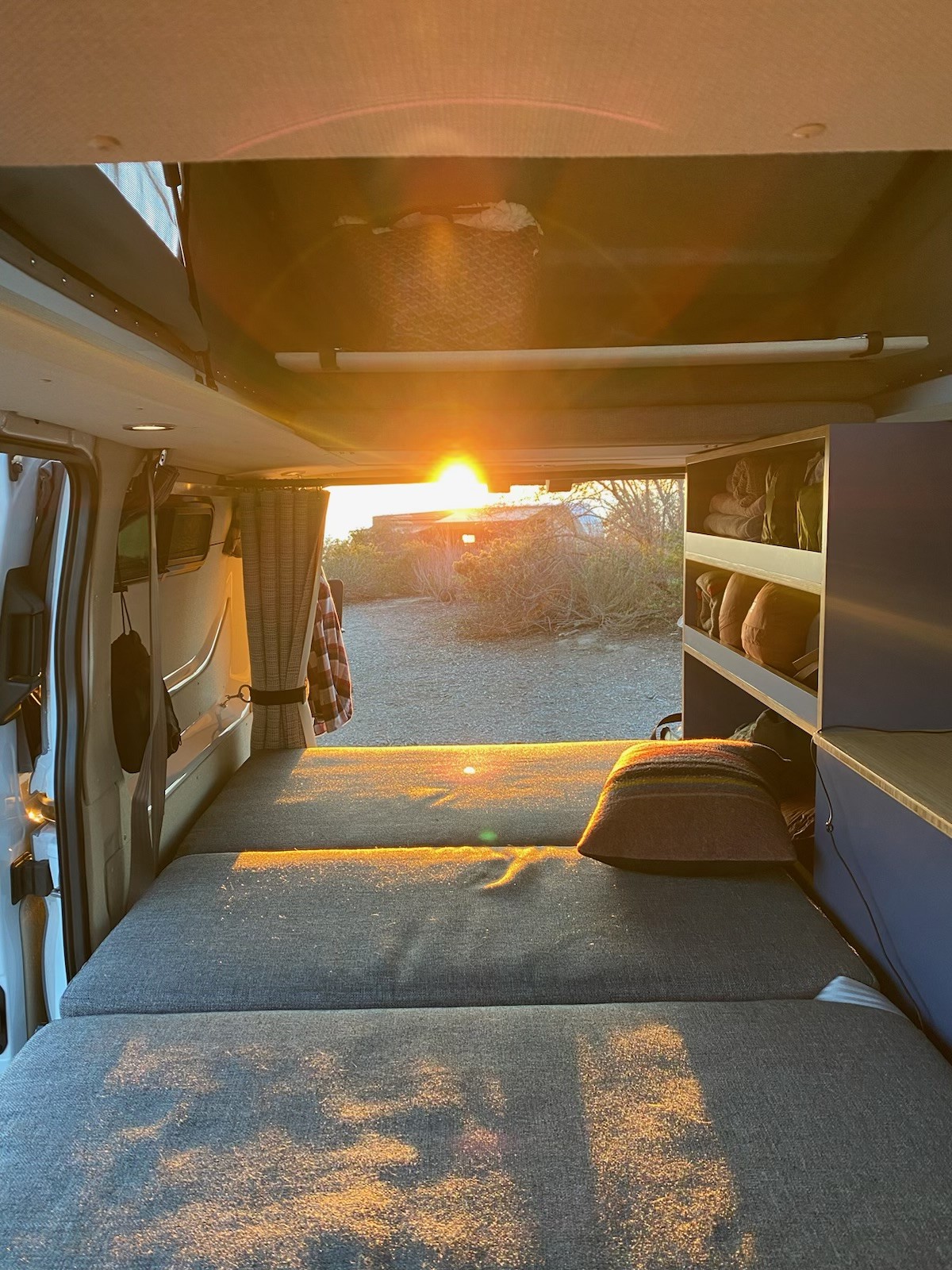 Golden light floods this cozy RV rental interior. Sunset over sparse landscape from van's open rear, with a bed & shelves.. Ford Transit 2022