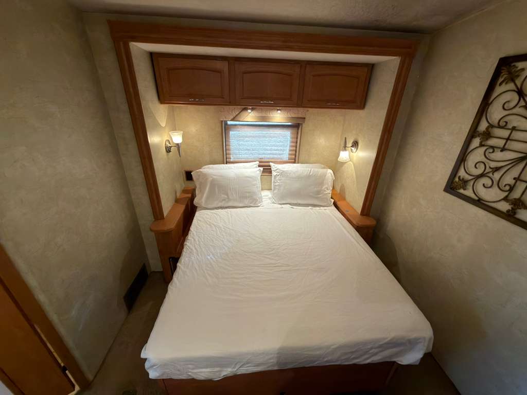 Experience comfort in this RV rental bedroom with a cozy bed, overhead cabinets, and warm lighting for your journey.. Winnebago Voyage 2008