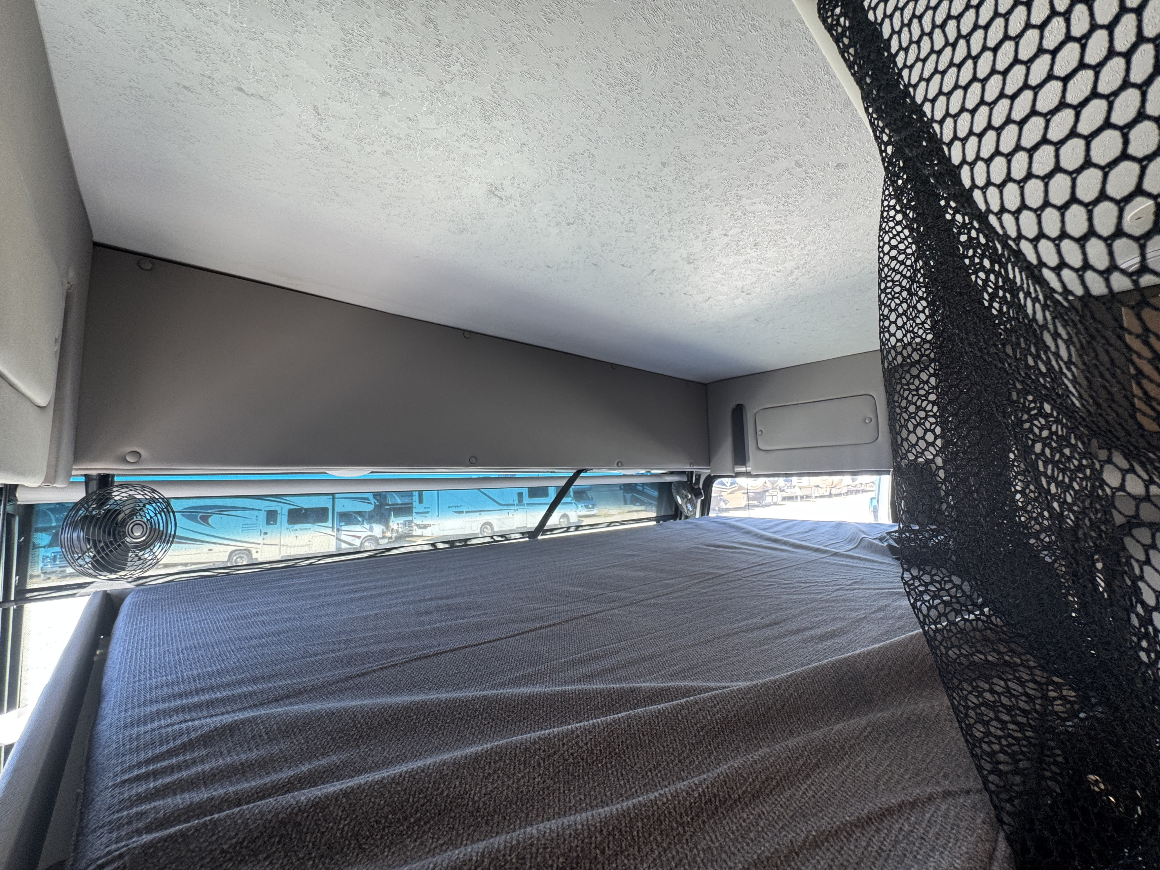 Inside an RV rental: a cozy cab-over bunk bed with fan, safety net, and a window view of other motorhomes.. Forest River Coachmen Encore 2022