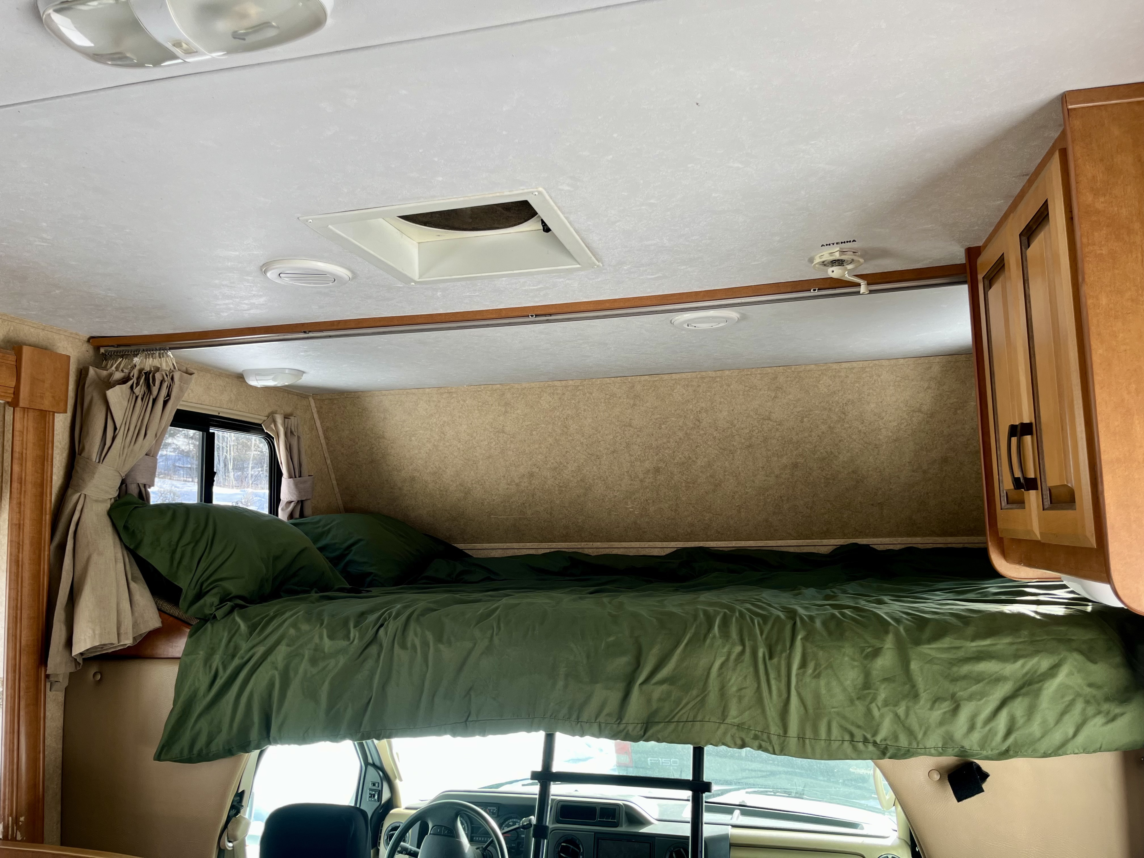 Inside an RV rental: a cozy cab-over bunk with green bedding. Features ceiling lights, vents, and wood cabinetry.. Forest River Sunseeker 2012