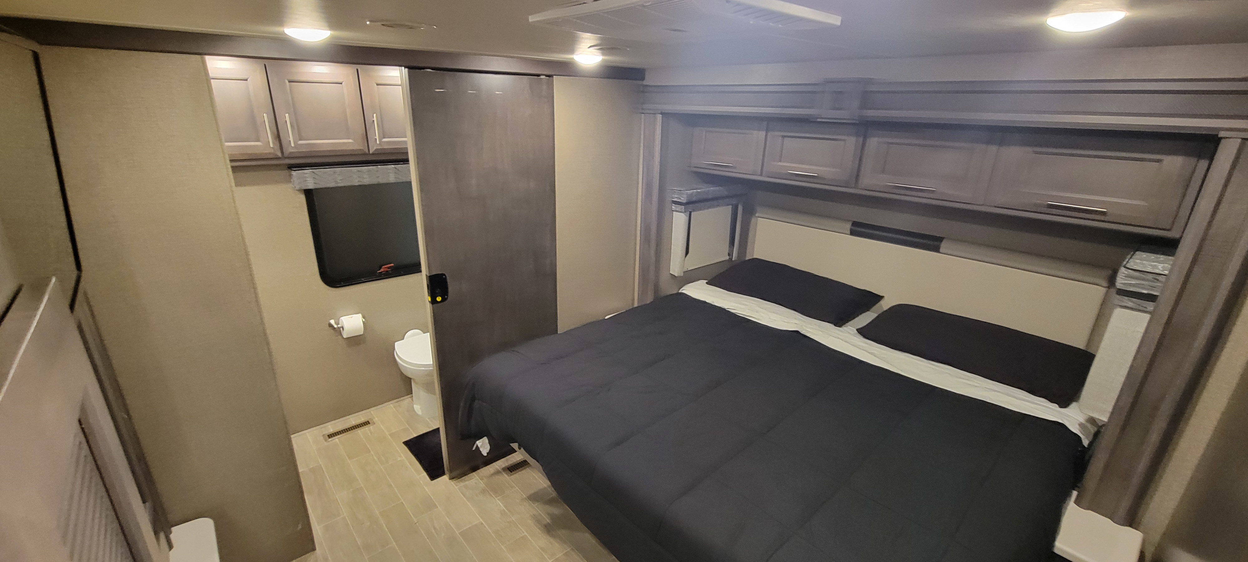 This RV rental bedroom features a black-covered bed, overhead cabinets, and a compact bathroom with a toilet behind a sliding door.. Thor Miramar 2022