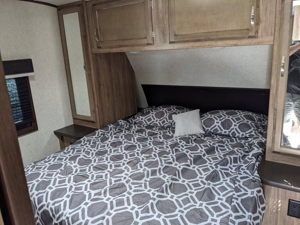 Inside your RV rental: a neat bedroom with a patterned bed, dark headboard, wooden cabinets, and mirrors.. Jayco Jay Feather 2019