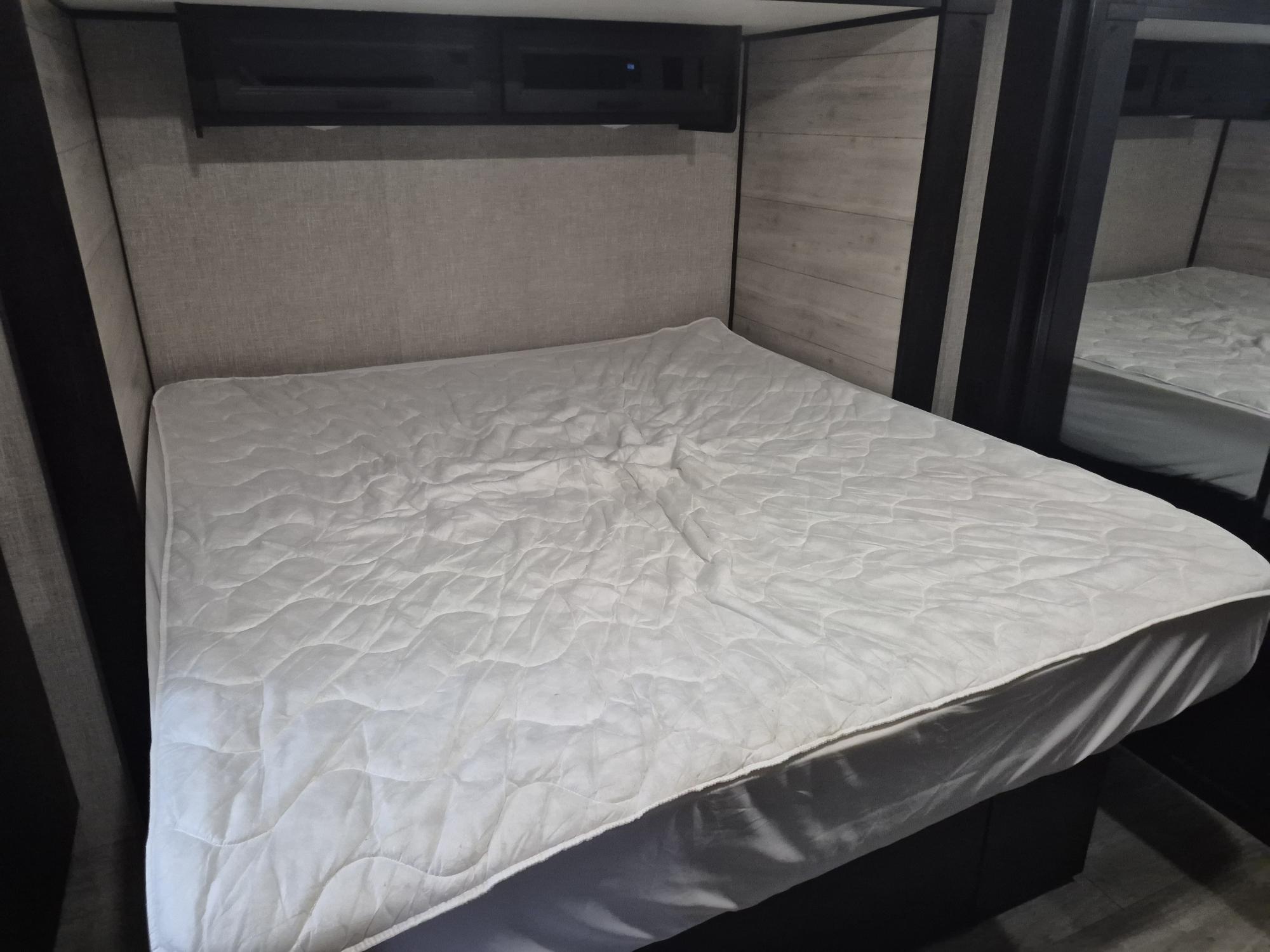 RV rental bed with a white quilted mattress, fabric headboard, and dark overhead storage in a compact space.. Jayco Jay Flight 2023