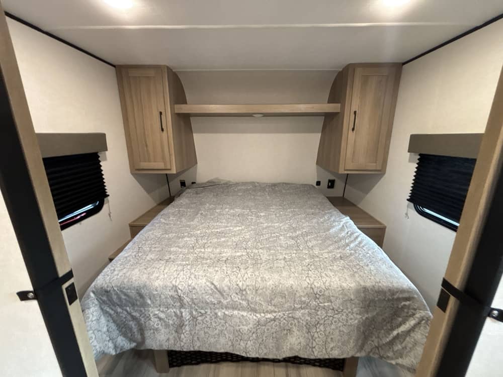 Cozy RV rental bedroom featuring a comfortable bed, overhead storage, and windows with black blinds for privacy.. Keystone RV Coleman Light 2025