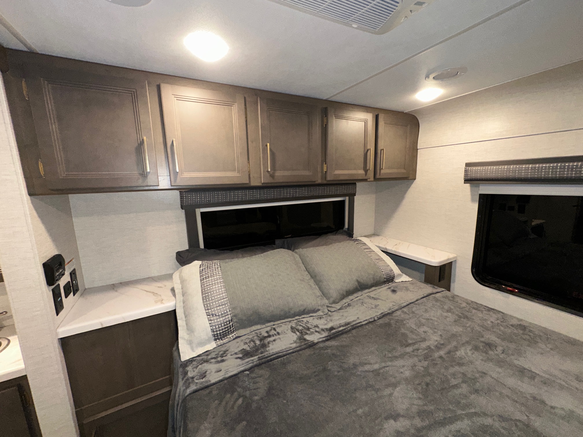 Cozy RV rental bedroom with a comfortable gray bed, overhead cabinets, and essential wall controls, offering practical travel comfort.. Thor Chateau 2024