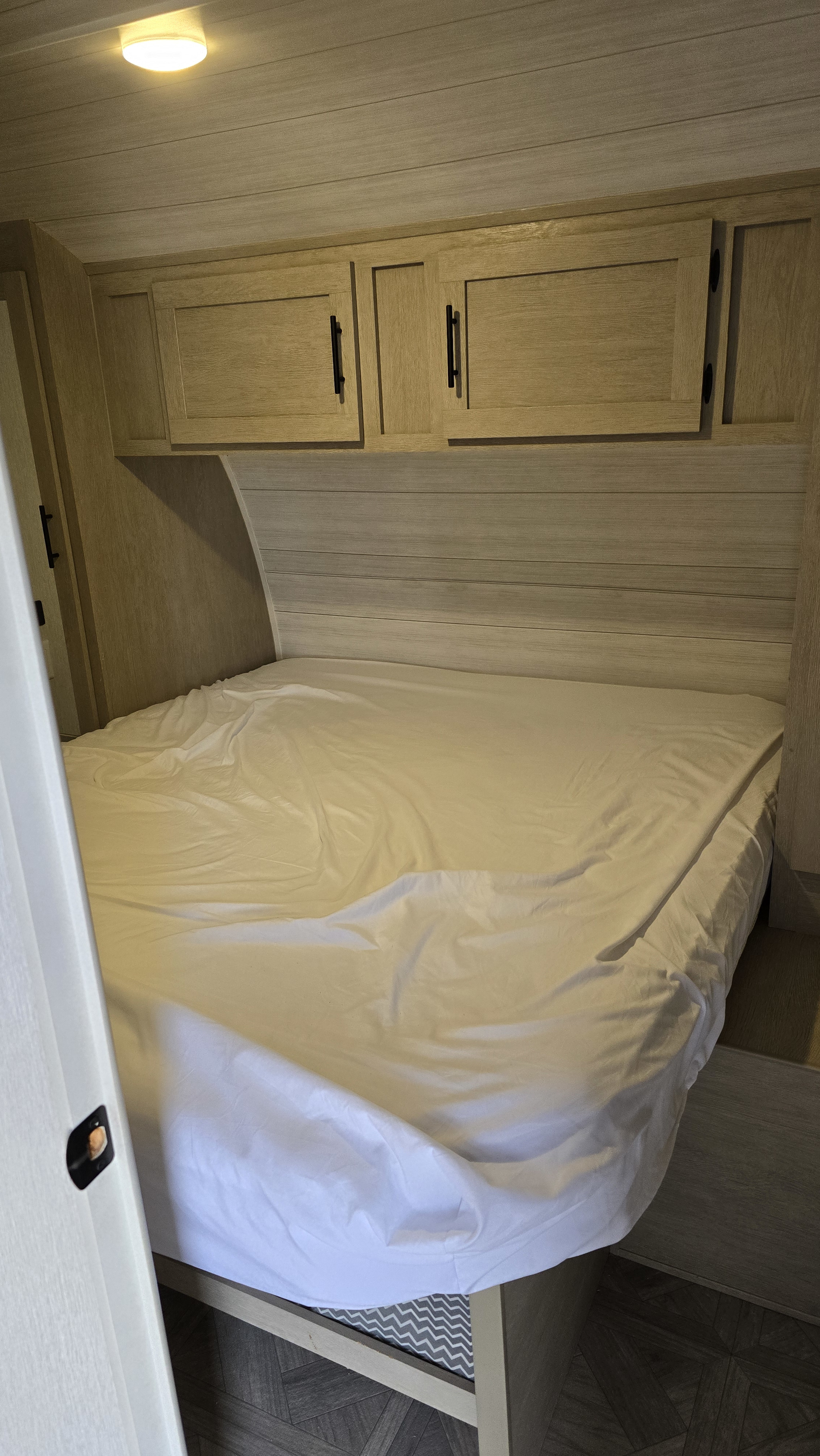 Cozy RV rental bedroom features a comfortable bed, wood paneling, overhead cabinets, and a bright ceiling light.. Forest River T27TDSS 2023