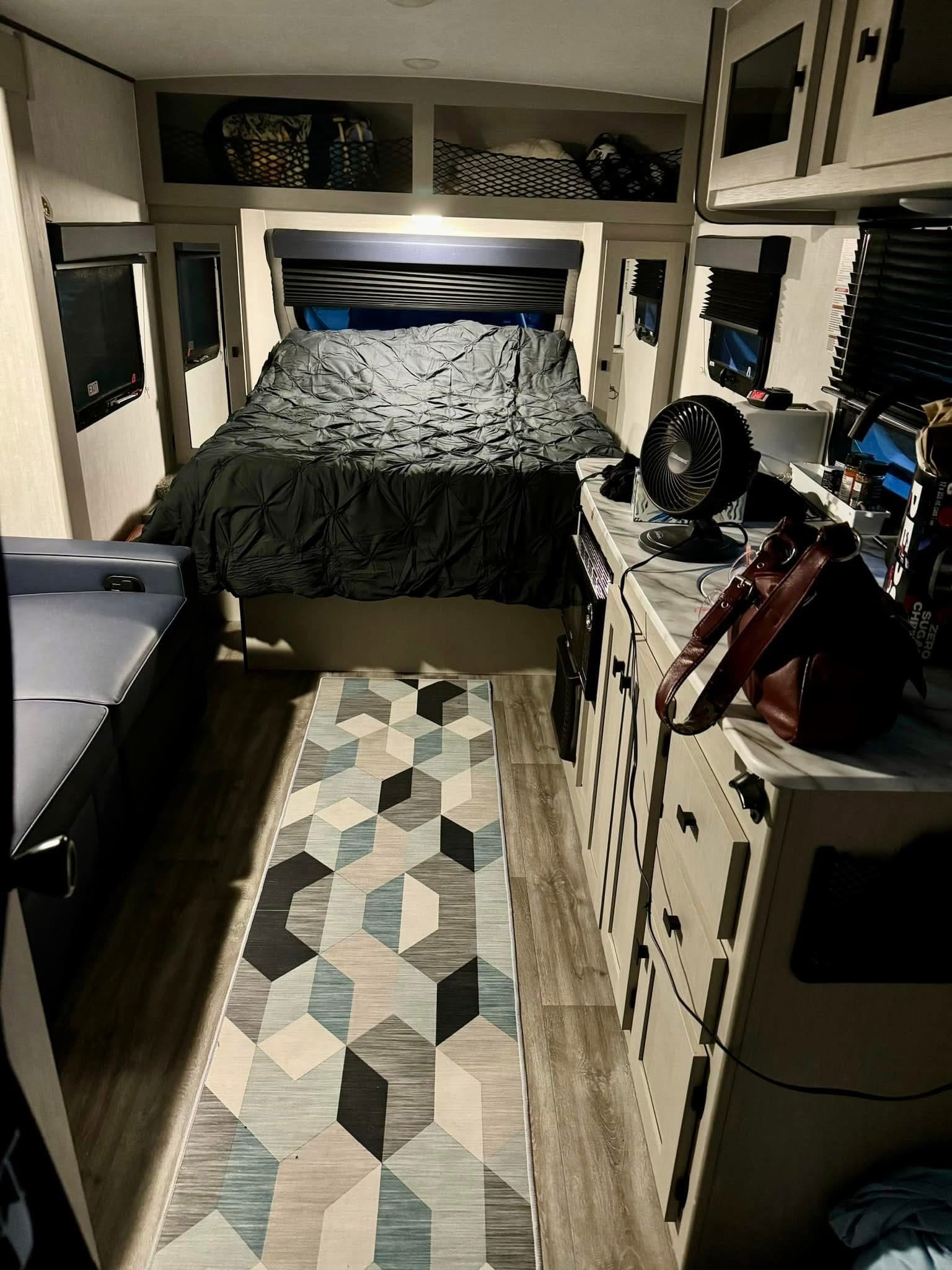 Rv rental bedroom: dark comforter on bed, geometric rug, light cabinets with a fan and bag, blue bench seating.. Coachmen Remote Off Grid 2024