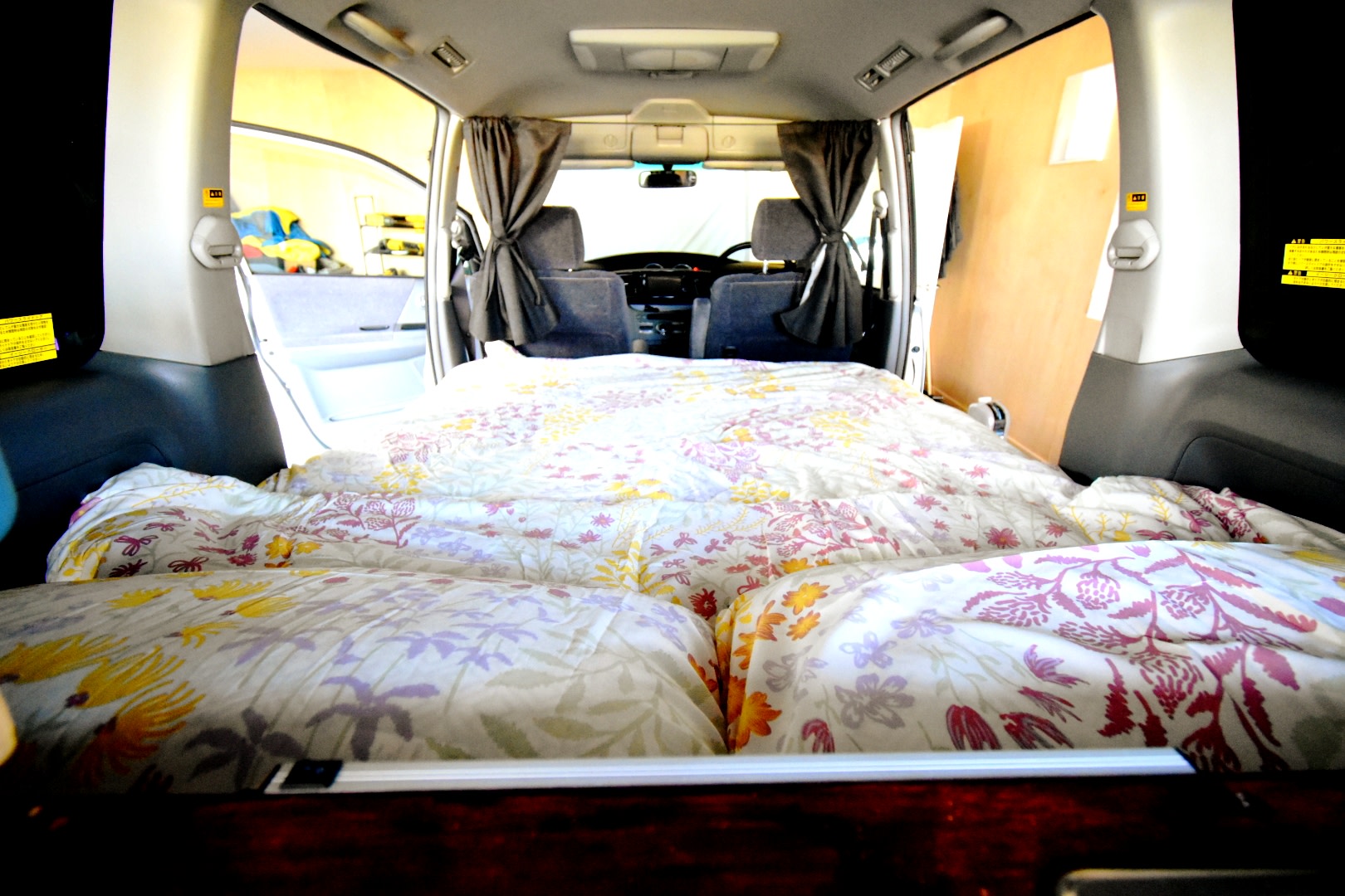 Cozy RV rental campervan interior with bright floral bedding, ready for travel. Front seats and dashboard visible, back prepared for sleeping.. Toyota Voxy 2005