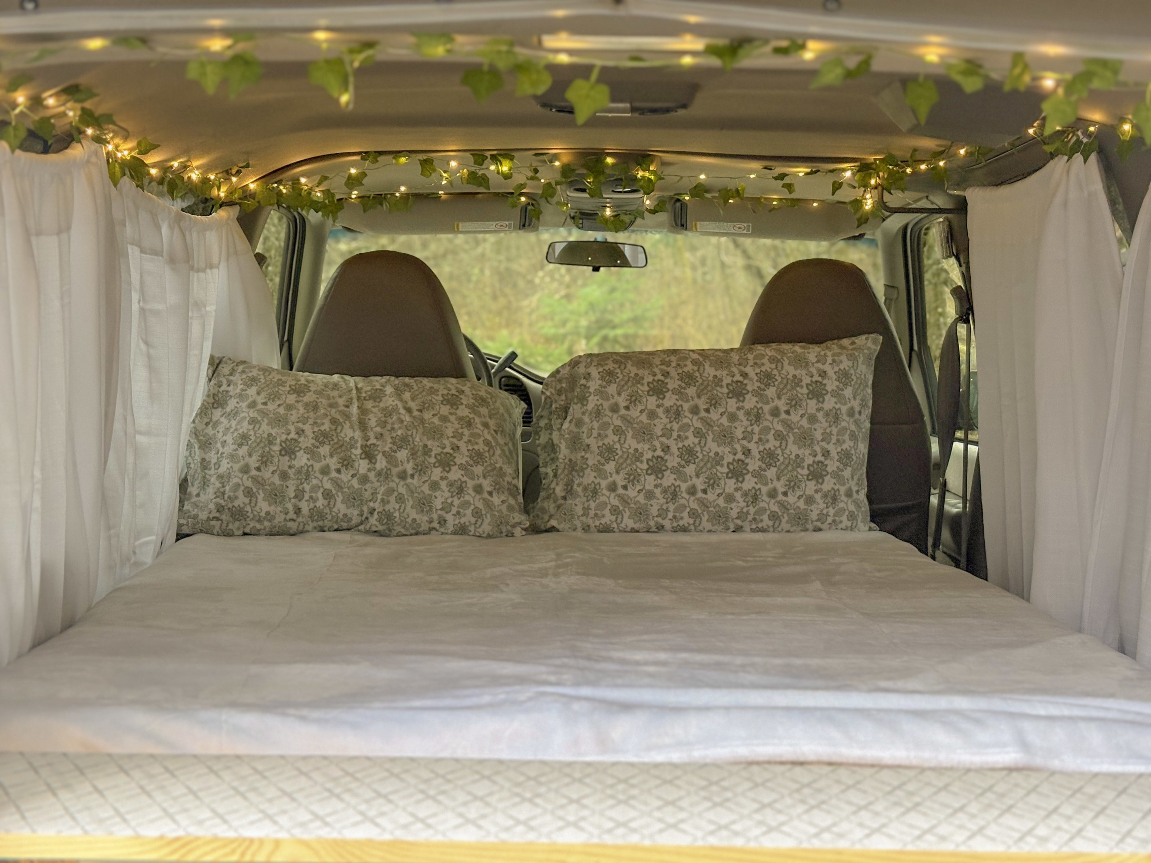 Cozy RV rental interior with a bed, patterned pillows, fairy lights, & curtains, offering a comfortable camping experience.. Chevrolet astro 2000