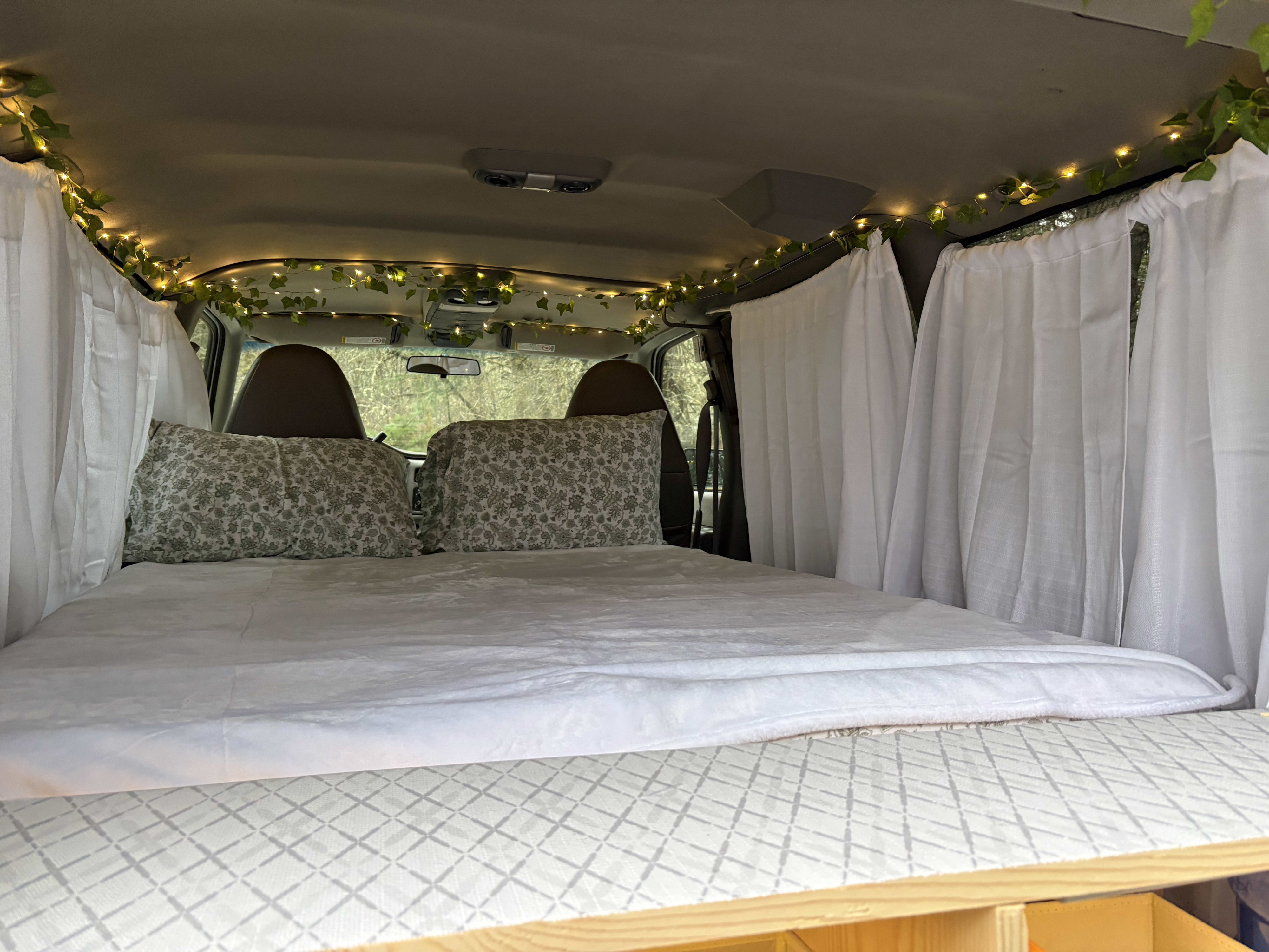 RV rental's cozy camper bed setup with patterned pillows, soft white bedding, fairy lights, & privacy curtains.. Chevrolet astro 2000