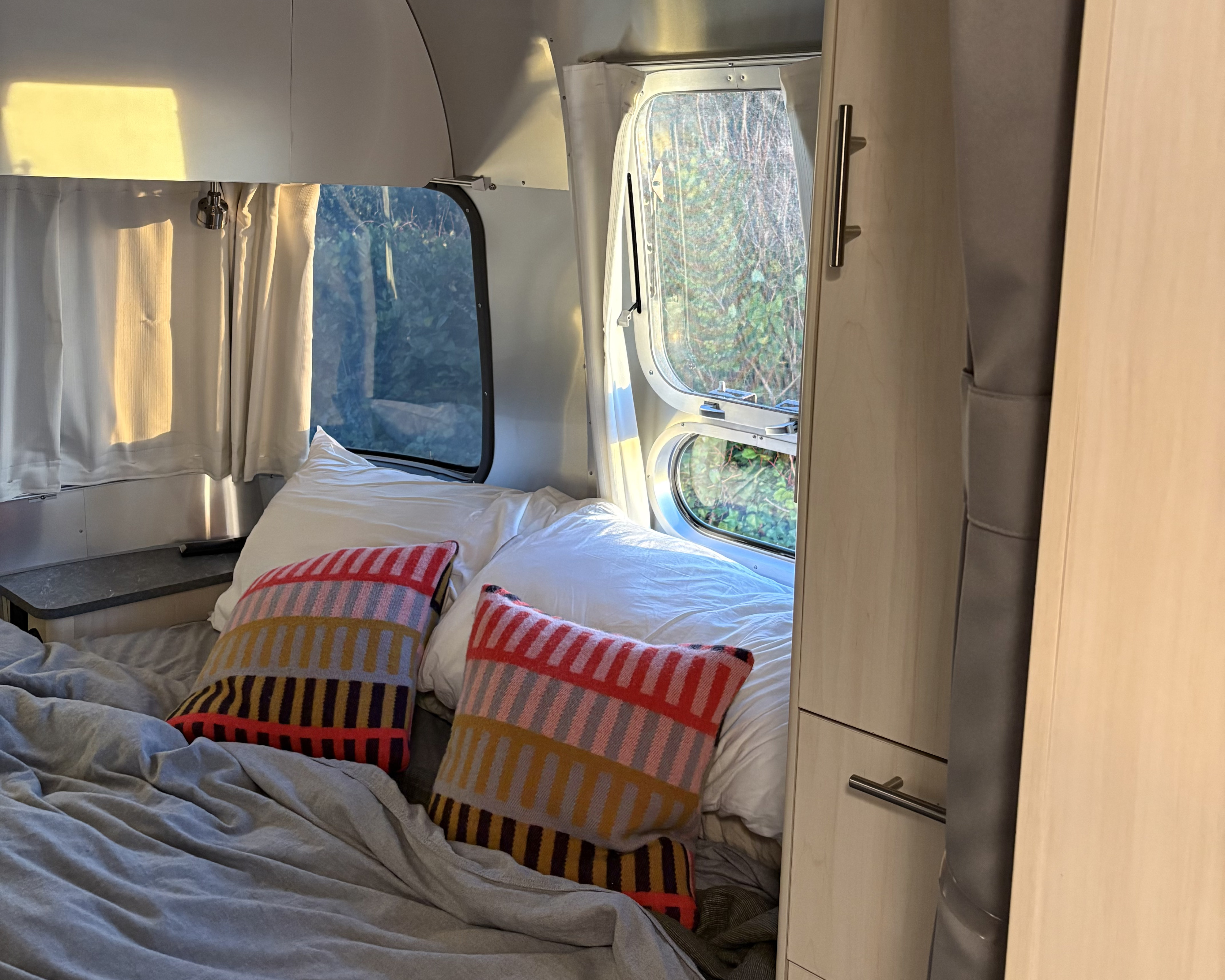 Sunlit rv rental Airstream bedroom: cozy white bedding, colorful pillows, and windows looking out to lush green nature.. Airstream Flying Cloud 2023