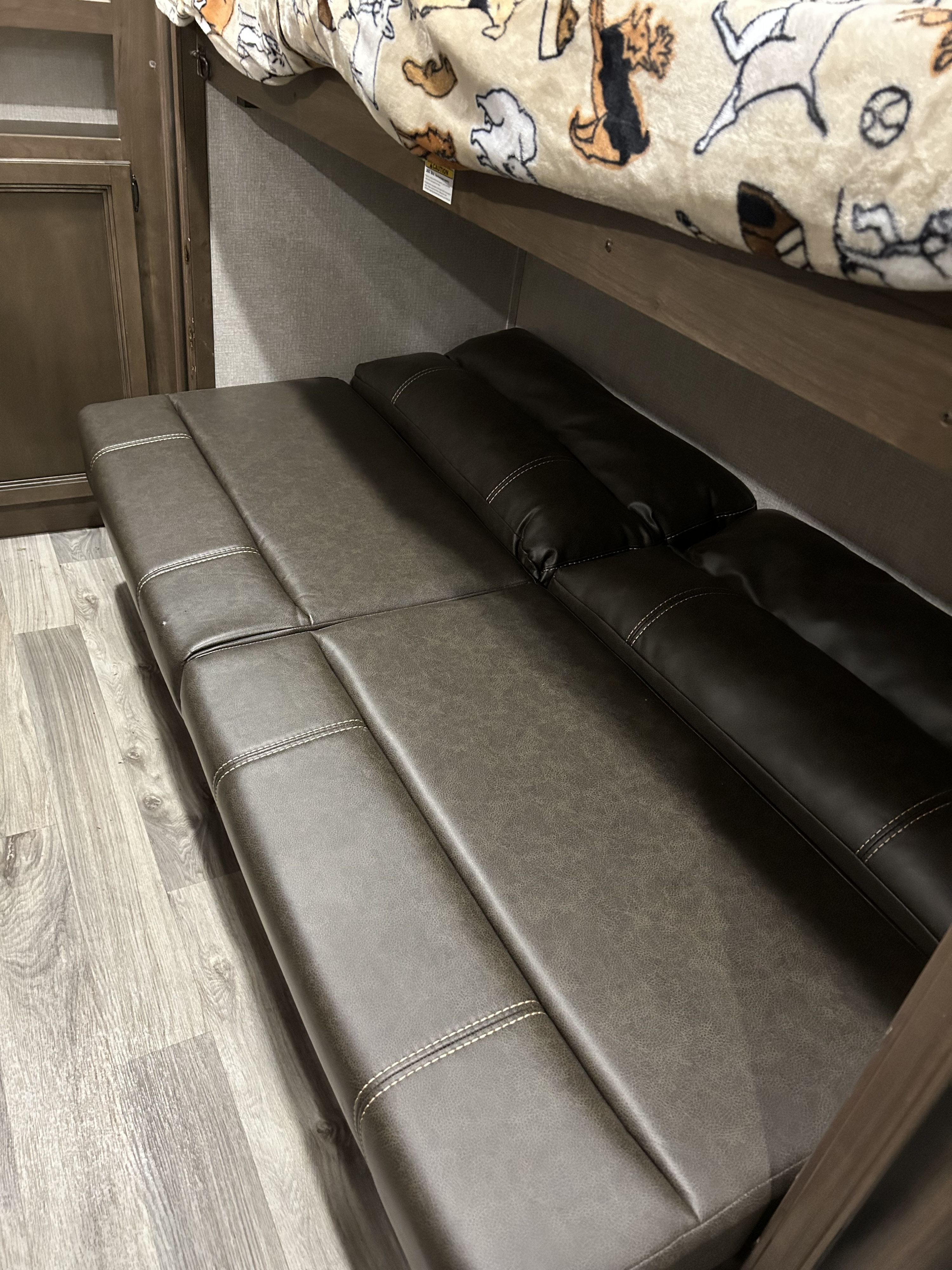 RV rental bunk bed with a dog-patterned blanket over a dark faux leather convertible sofa and wood flooring.. Keystone RV Cougar 2021