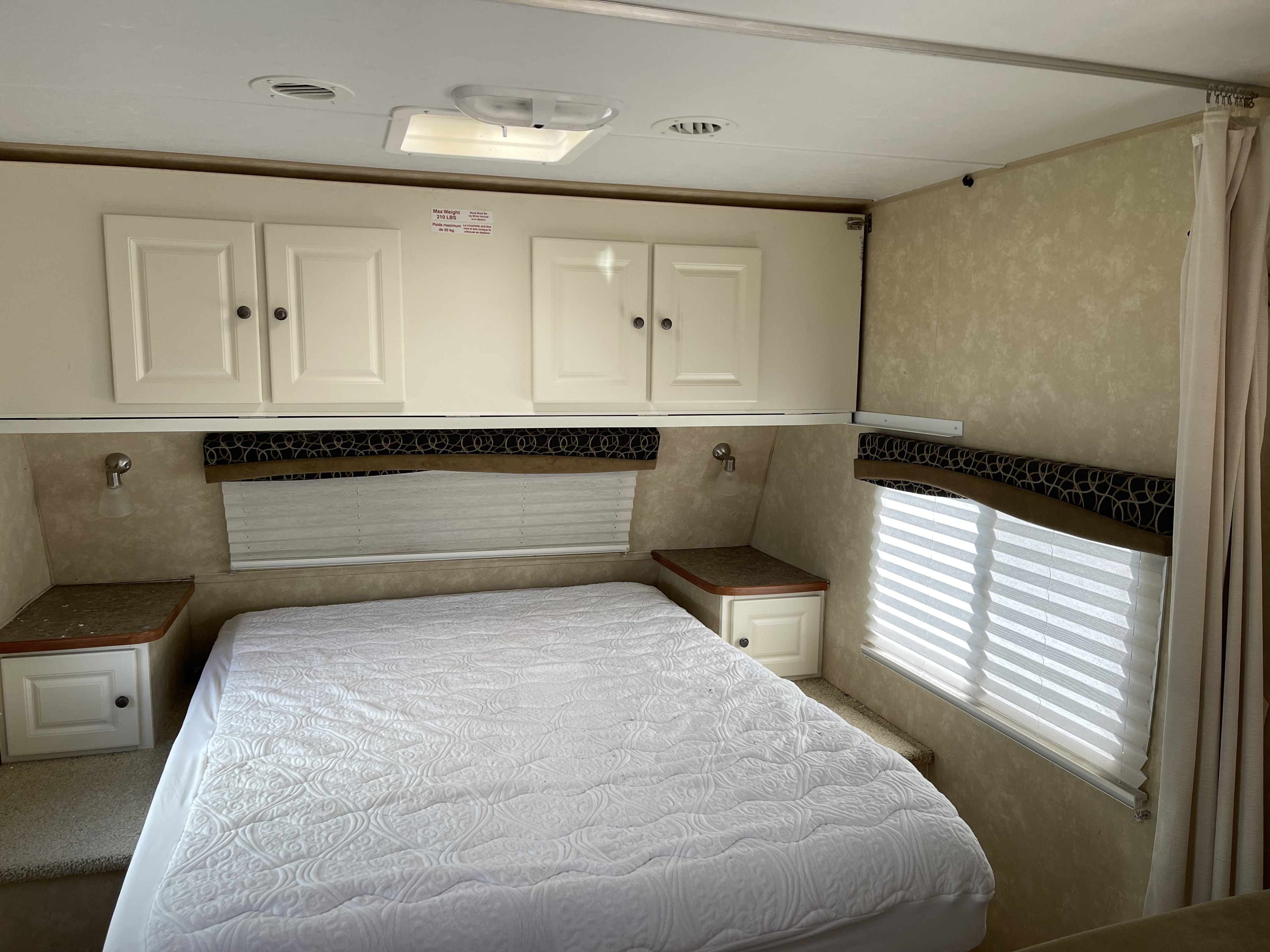Cozy RV rental bedroom featuring a clean white bed, overhead storage cabinets, nightstands, and windows with blinds.. Cruiser RV Fun Finder 2011