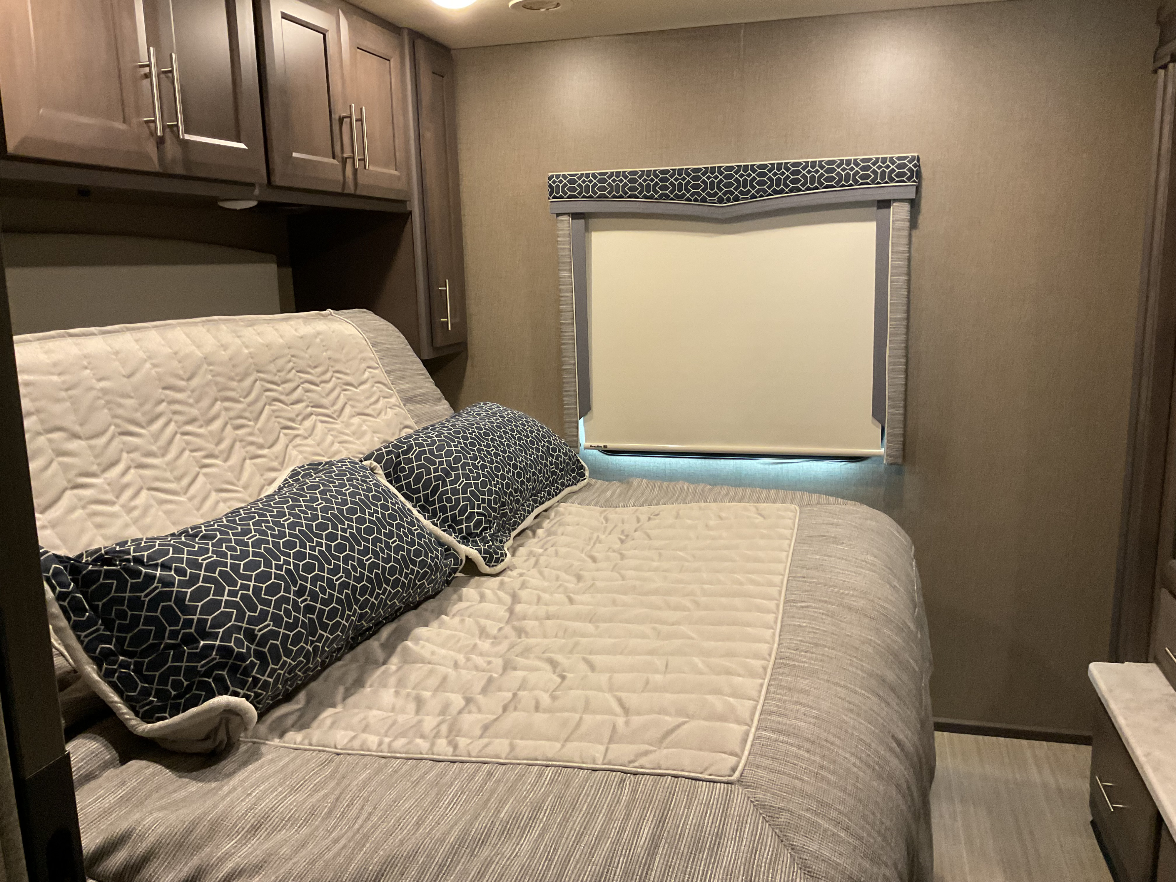 Cozy RV rental bedroom featuring a striped bed with patterned pillows, dark wood cabinets, and a window with stylish valance.. Thor Magnitude 2022