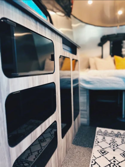 View inside an RV rental: light wood cabinet with dark cutouts, cozy bed with yellow pillows, & patterned rug.. Airstream Basecamp 2022