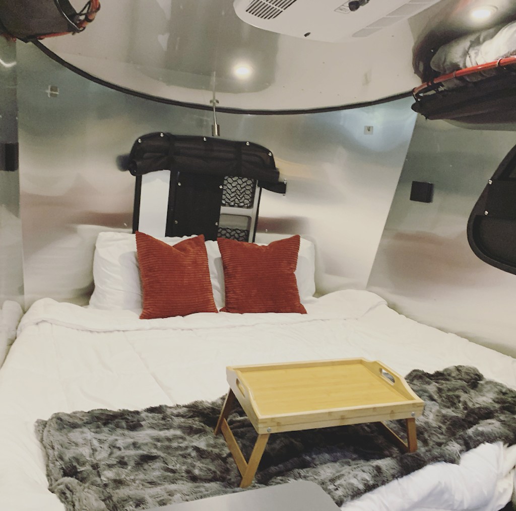 Inside a comfortable RV rental: a bed with white bedding, orange pillows, and a wooden tray on a faux fur throw. Cozy bedroom.. Airstream Basecamp 2022
