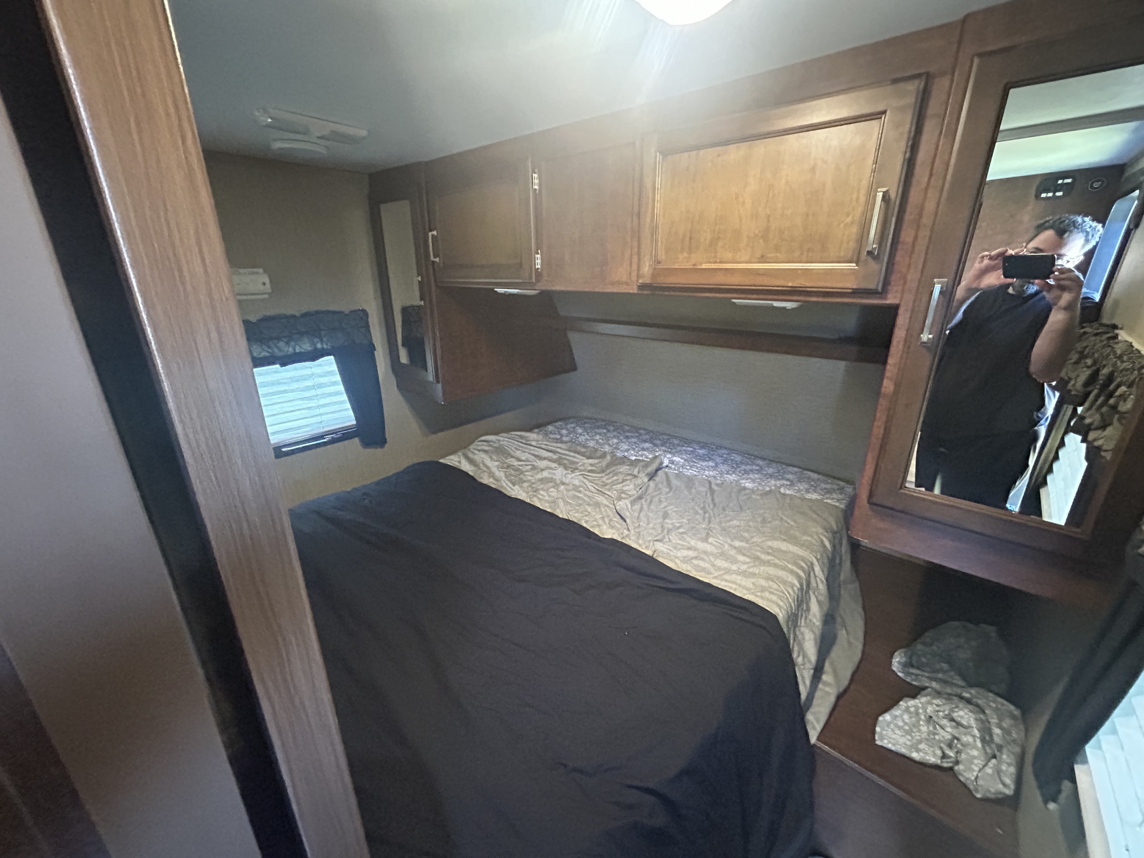 Compact RV rental bedroom with a bed, overhead wood cabinets, and a window with blinds. A person is reflected in the mirror.. Keystone RV Outback Ultra-Lite 2017