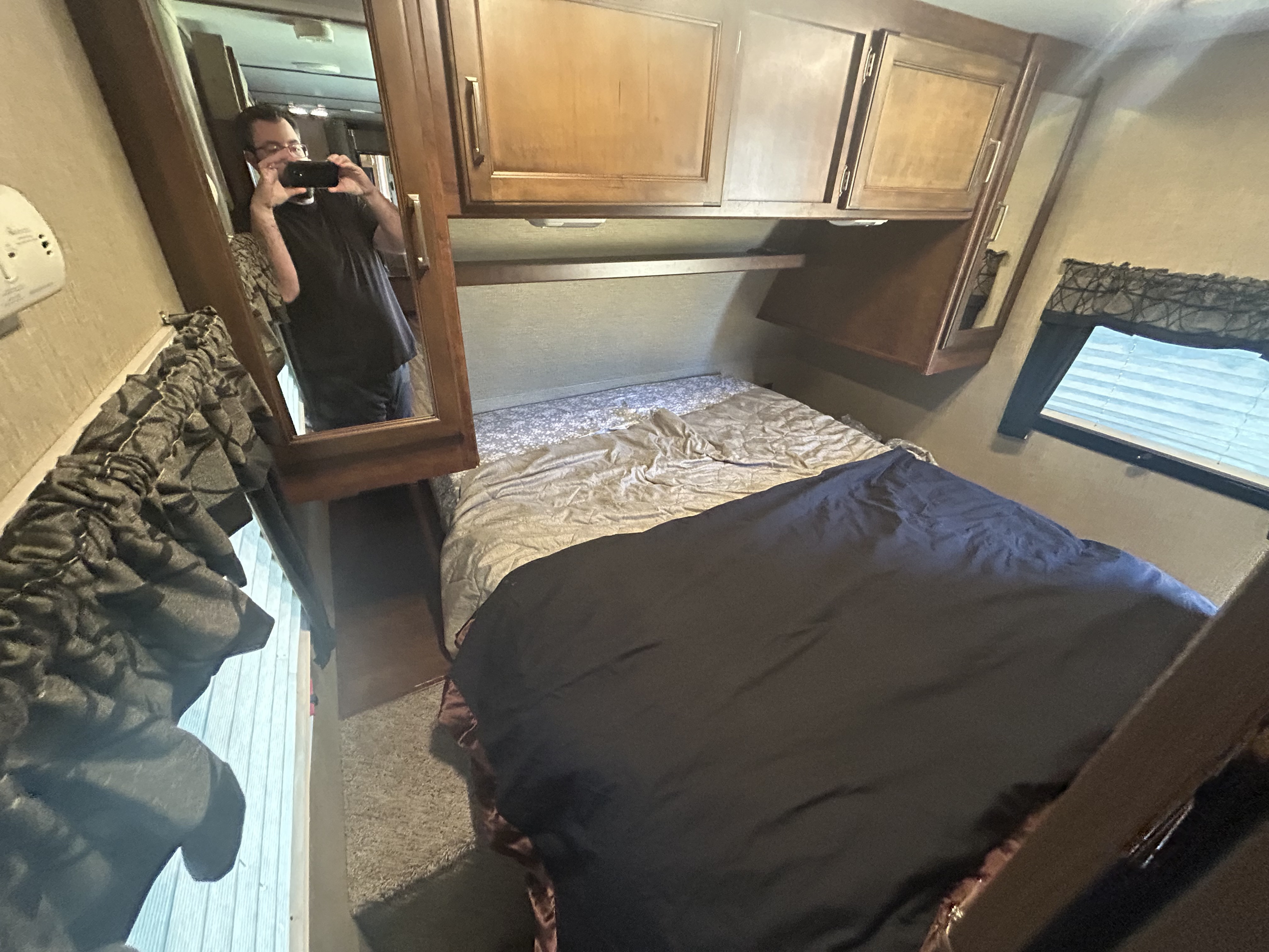 Inside an RV rental bedroom: a bed with dark bedding, mirrored closet reflecting the photographer, overhead cabinets, and a window.. Keystone RV Outback Ultra-Lite 2017