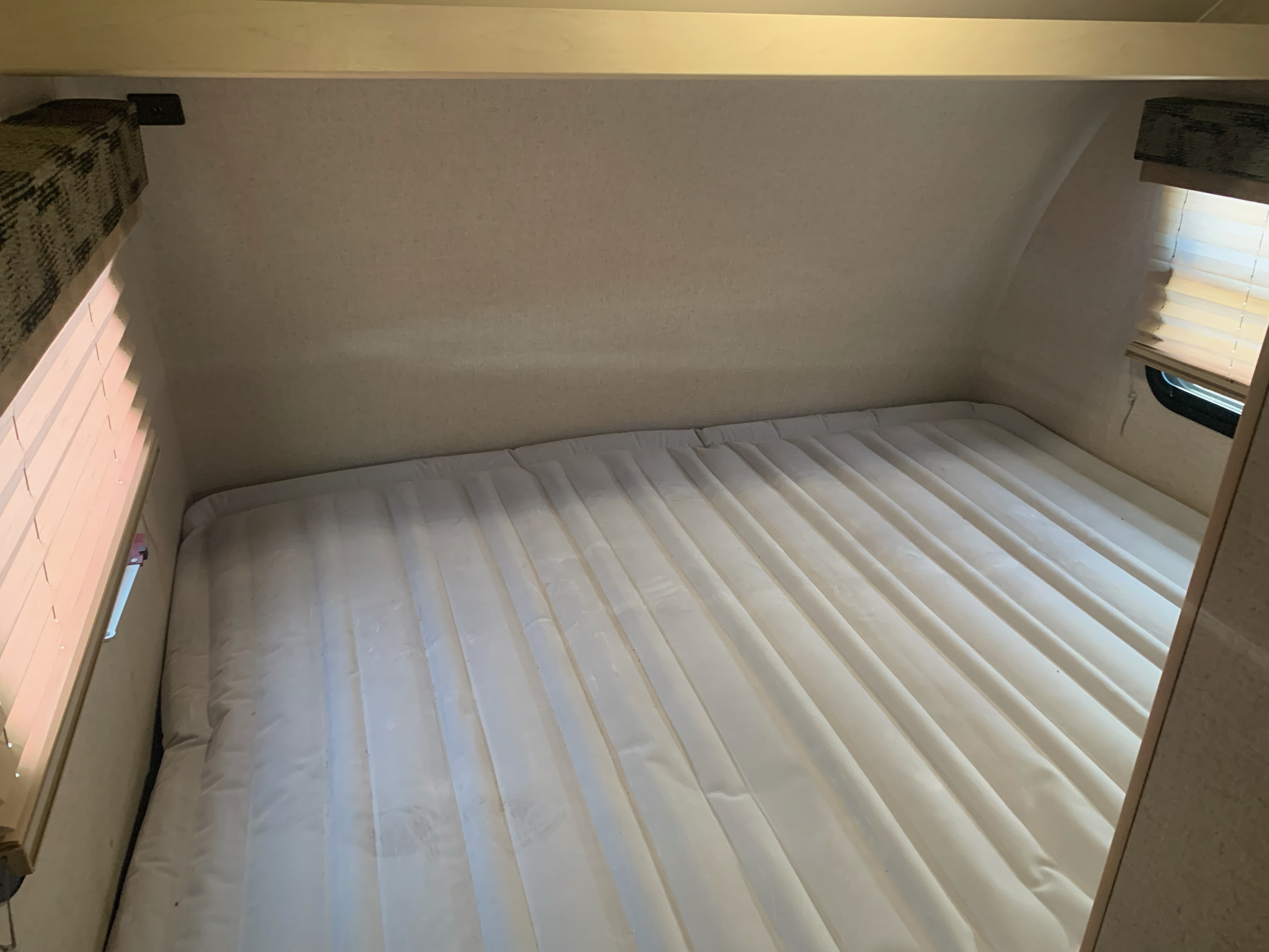 RV rental interior showing a compact sleeping area with a mattress, sloped ceiling, and window blinds.. Winnie Drop Winnie Drop Trailer 2018