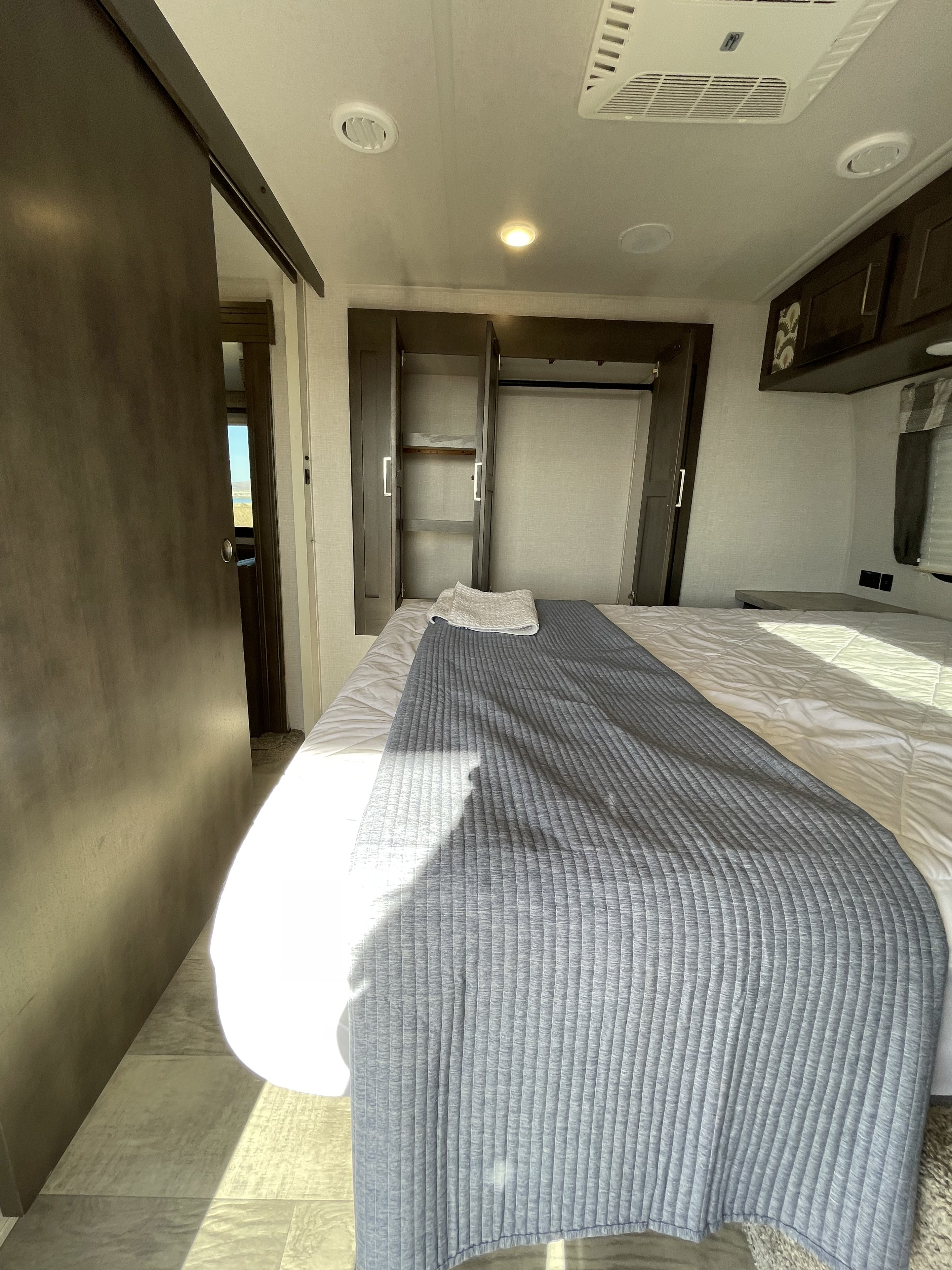 Bright RV rental bedroom: Features a bed with blue and white bedding, open closet, and a wooden sliding door, bathed in sunlight.. Forest River Flagstaff Super Lite 2021