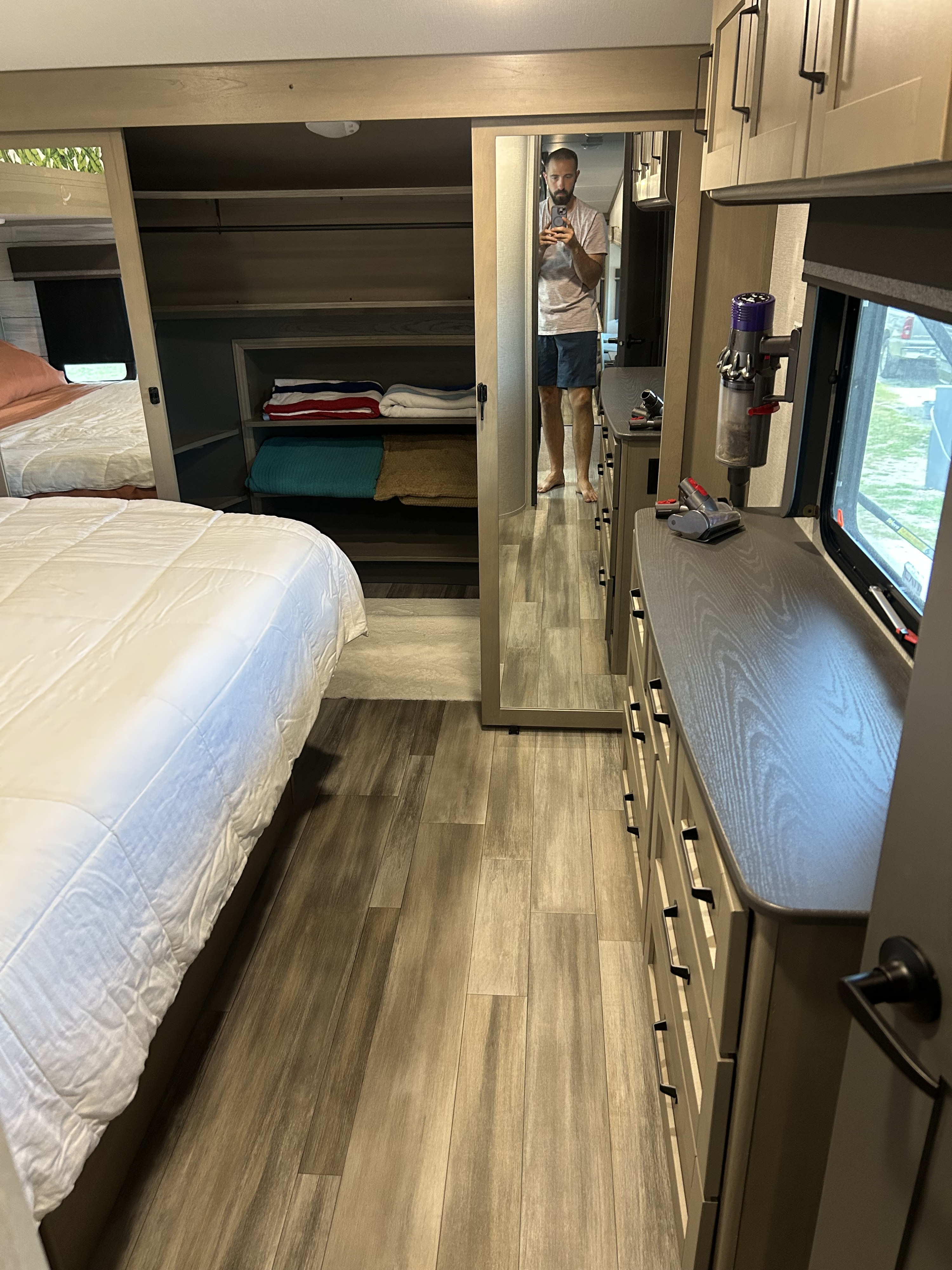 RV rental bedroom with light wood-look floor, large bed, modern dresser, and built-in storage. Photographer reflected in mirror.. Grand Design Reflection 2023