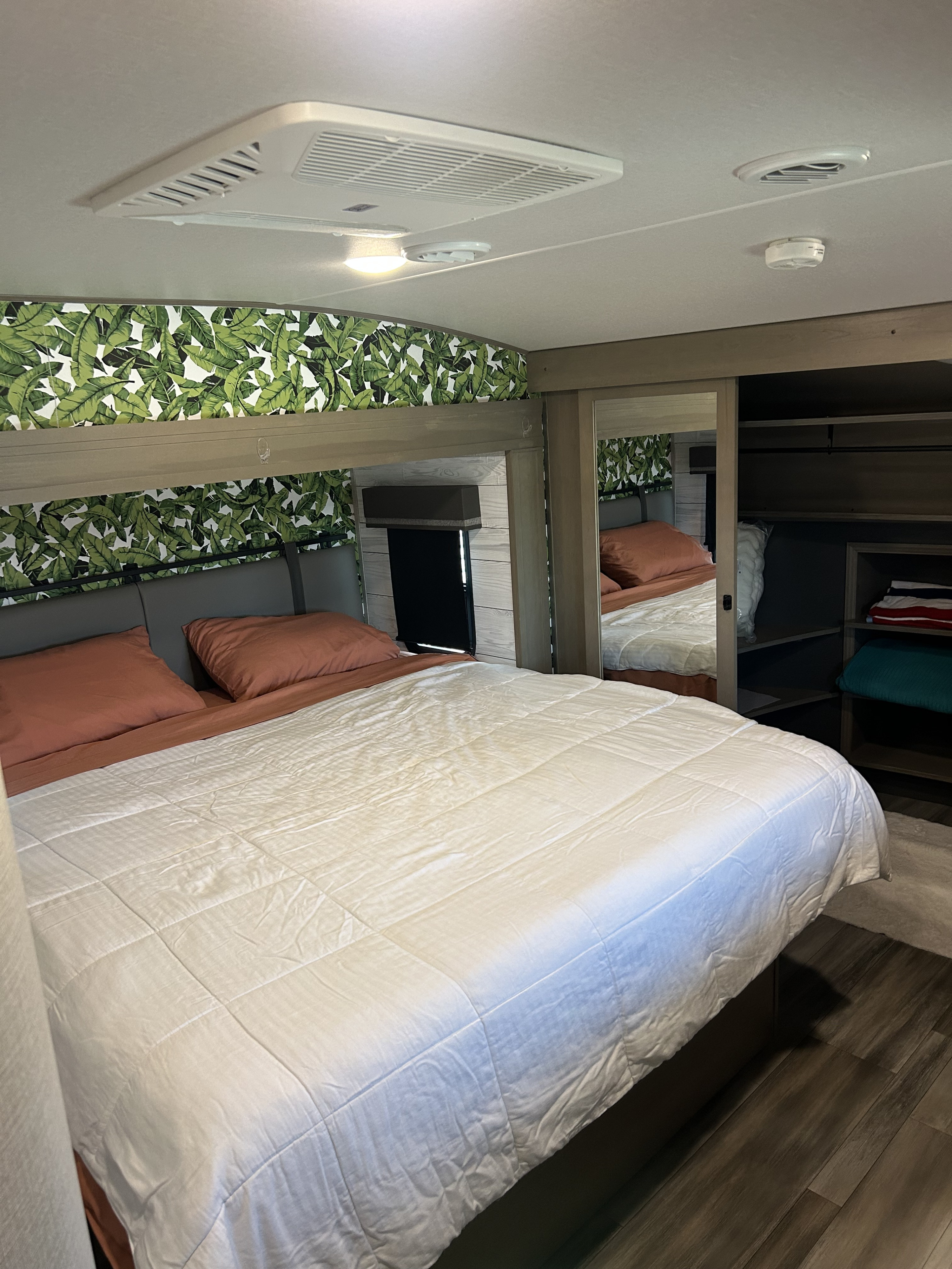 RV rental bedroom with a large bed, tropical leaf wallpaper, overhead AC unit, and a built-in mirrored closet.. Grand Design Reflection 2023