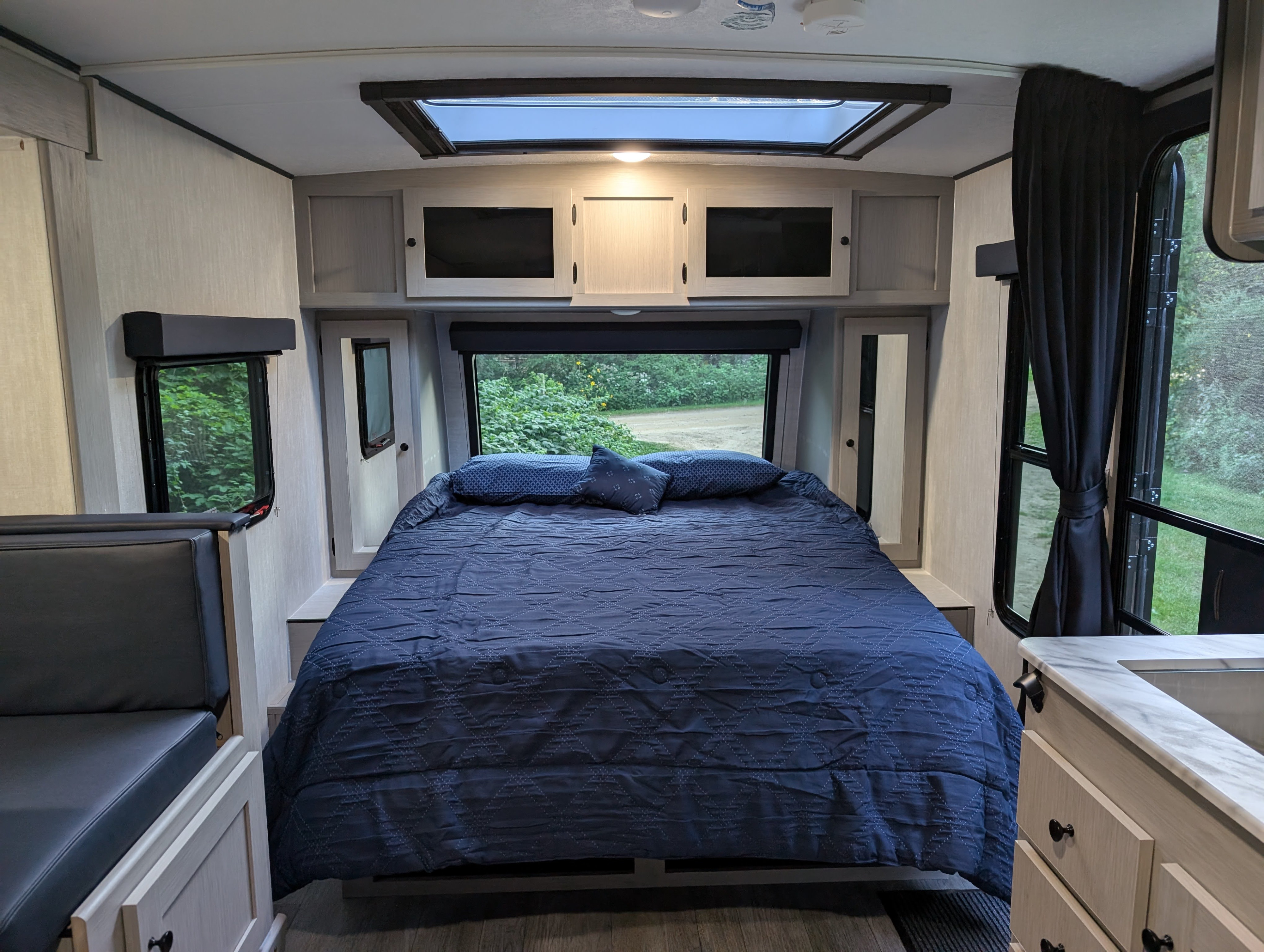 Modern rv rental interior with a comfy blue bed, skylight, dinette, and outdoor views of greenery.. Coachmen Apex Nano 2025