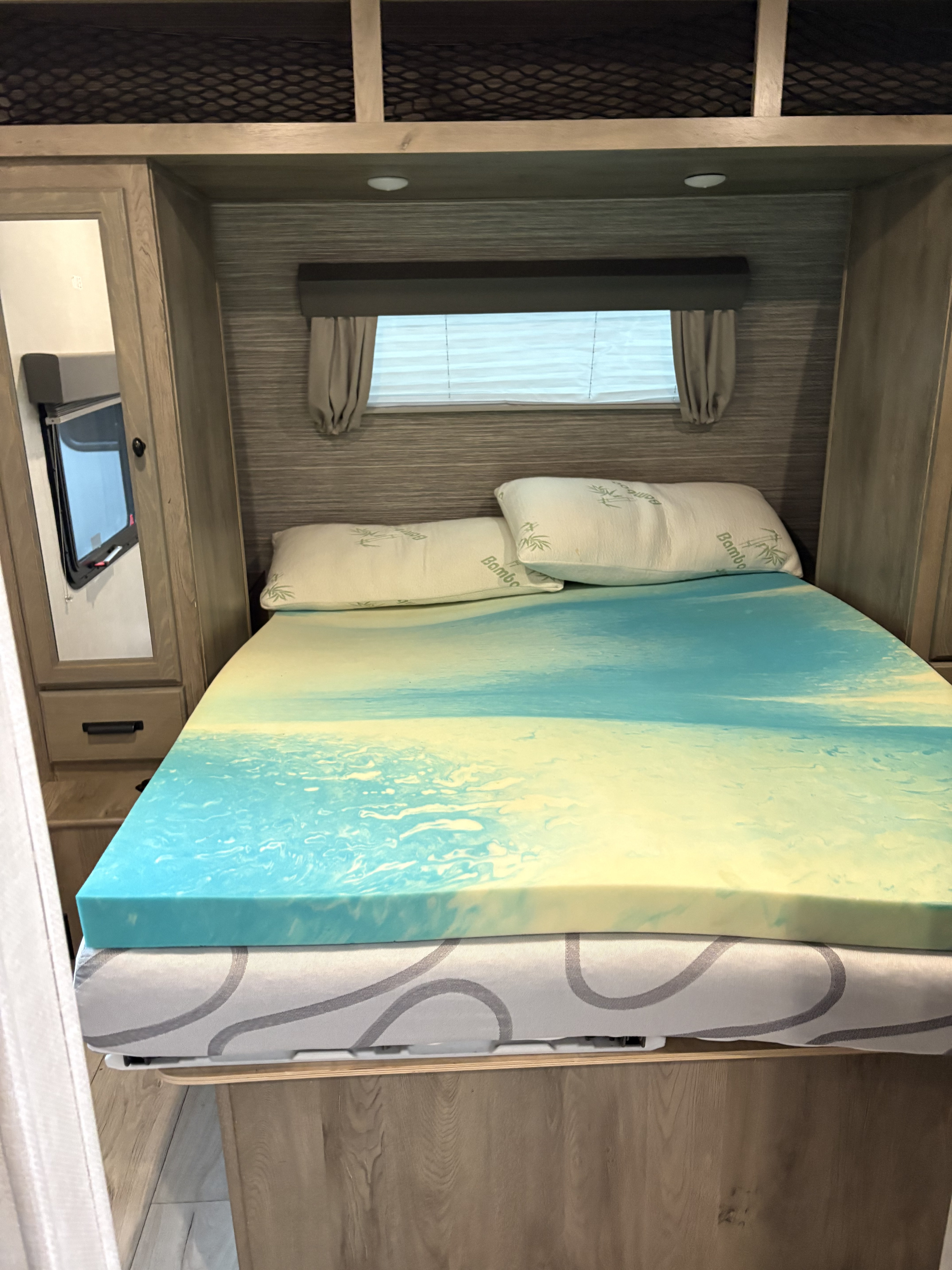Cozy RV rental bedroom featuring a bed with a blue & yellow foam topper, Bamboo pillows, window, and wood cabinetry.. Forest River Surveyor Legend 2022