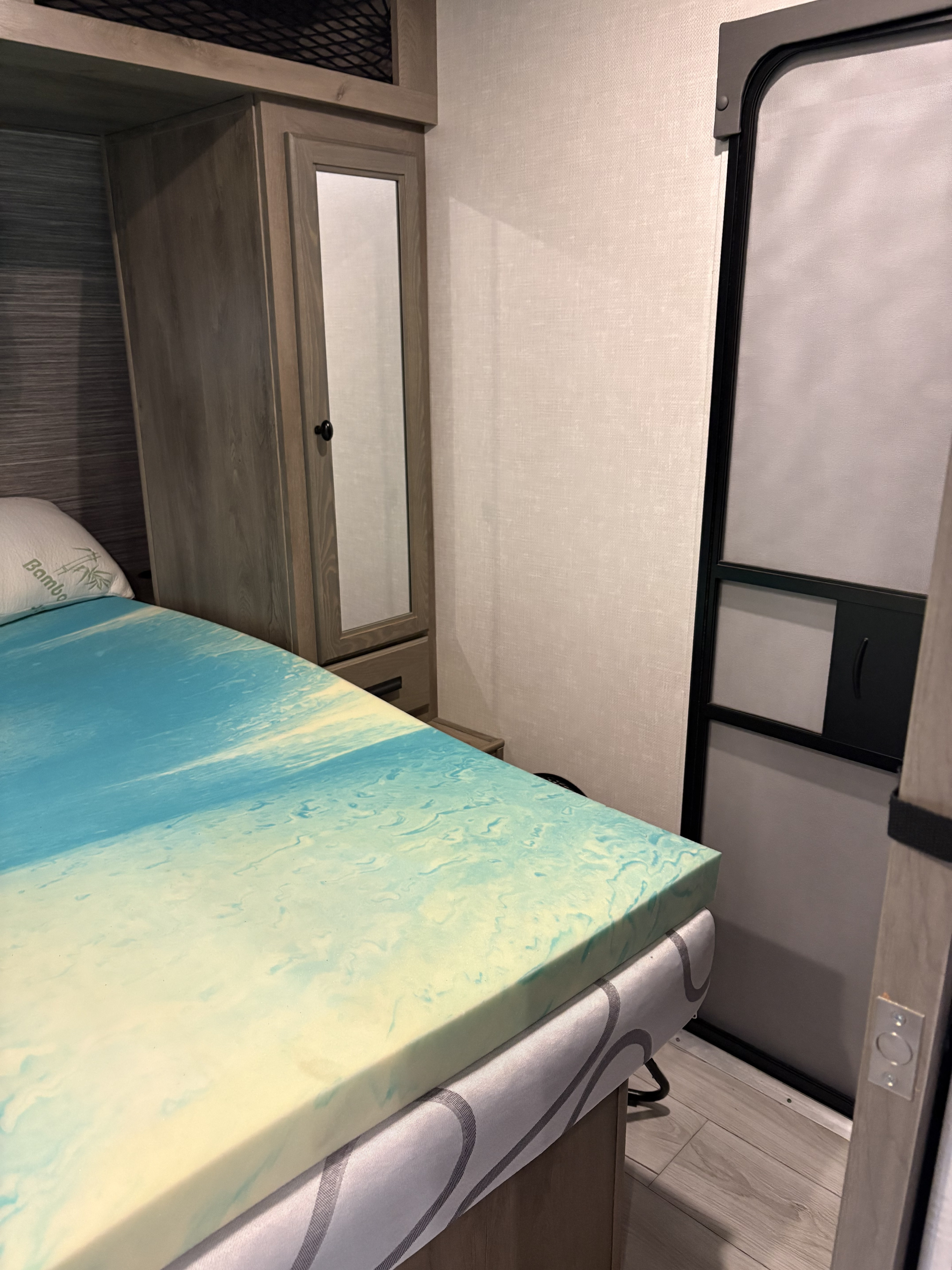 An RV rental bedroom interior with a bed featuring a blue/yellow memory foam mattress topper, mirrored wardrobe, and screen door.. Forest River Surveyor Legend 2022