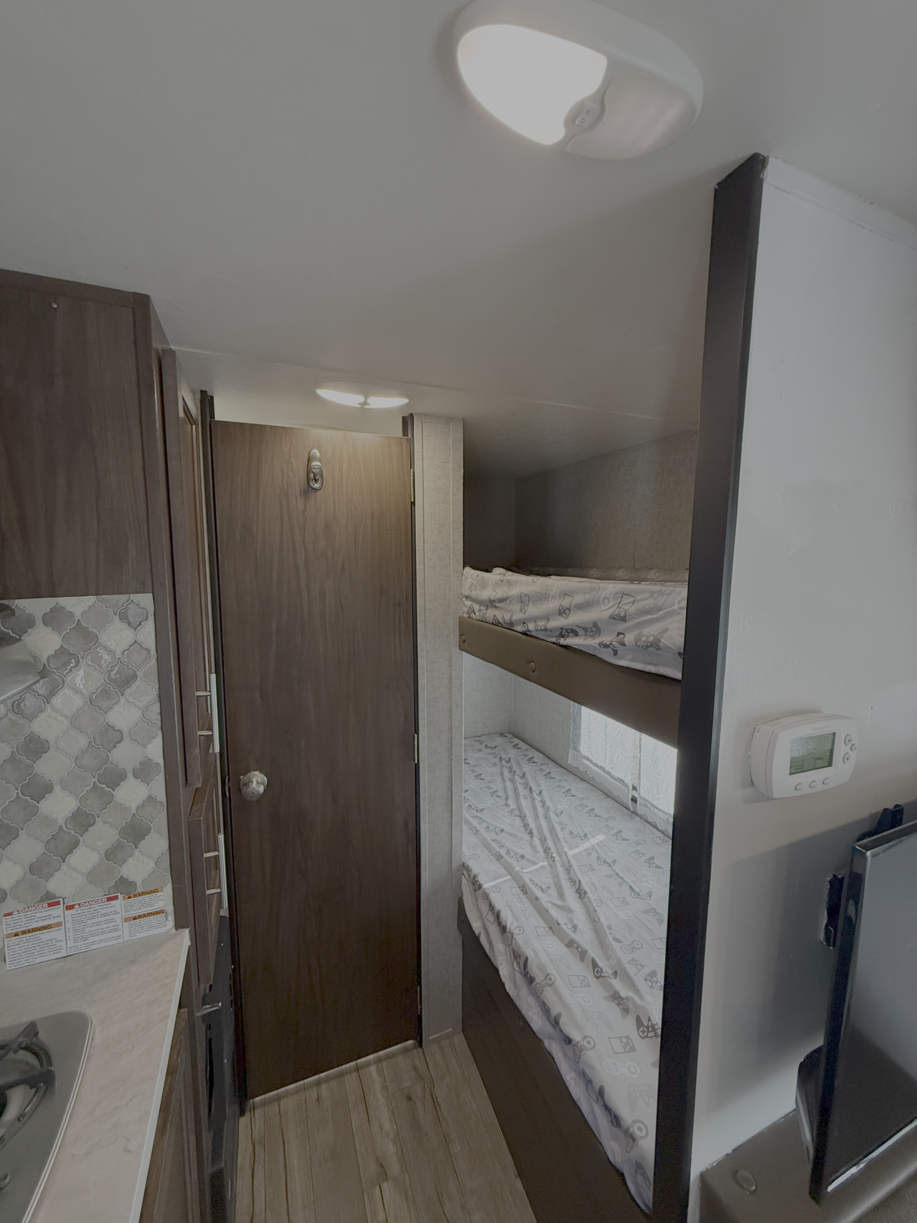 This RV rental interior shows bunk beds with patterned sheets, a small kitchenette, a door, and a wall-mounted thermostat.. Forest River Cherokee Wolf Pup 2018
