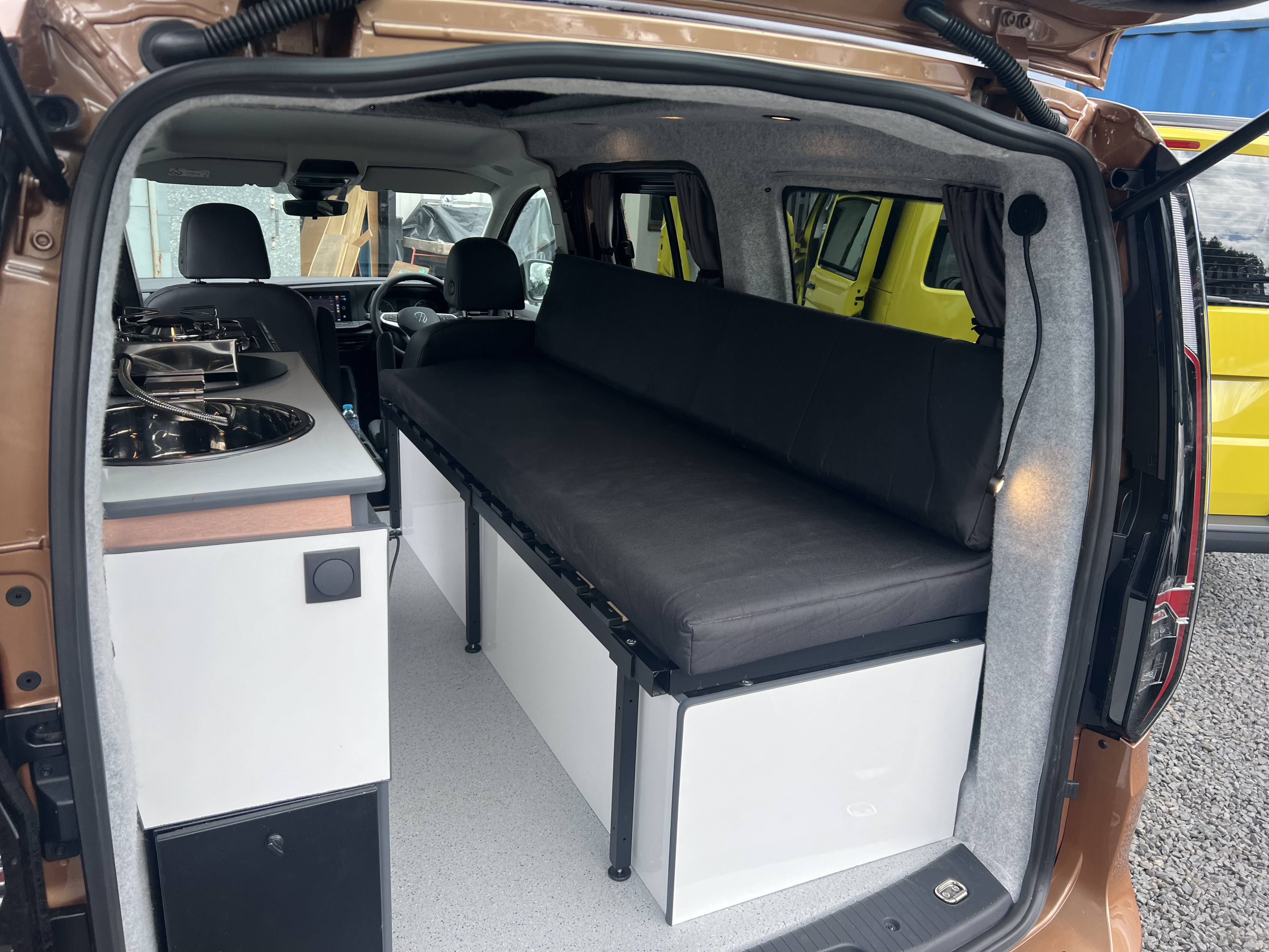 Compact **rv rental** campervan interior showcasing a kitchenette with a sink and stove, and a convertible bed with storage.. Volkswagen Caddy Maxi 2023