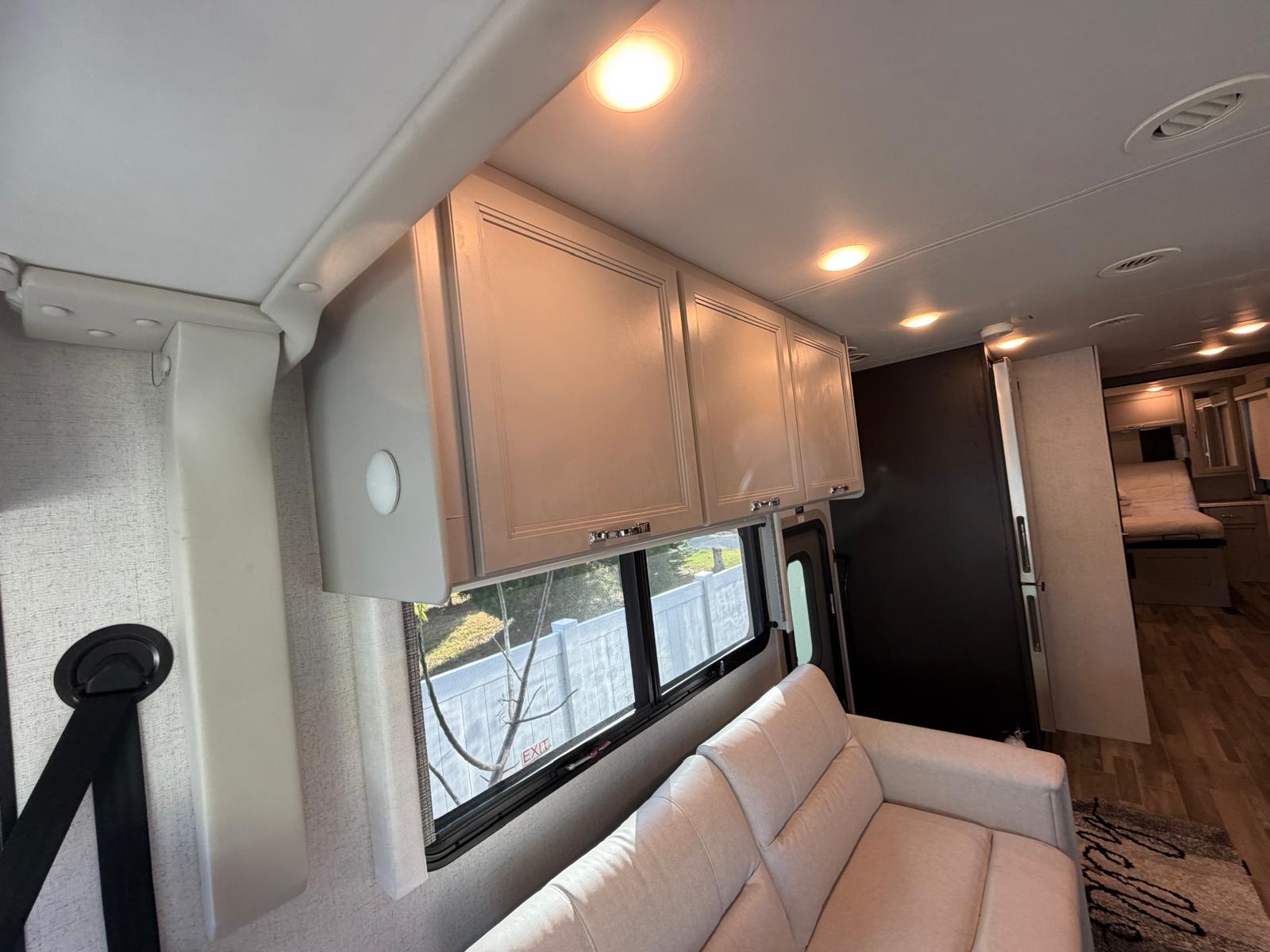 Bright RV rental living space with a comfy sofa, overhead cabinets, and windows. A bedroom area is visible further back.. Thor Palazzo 2024