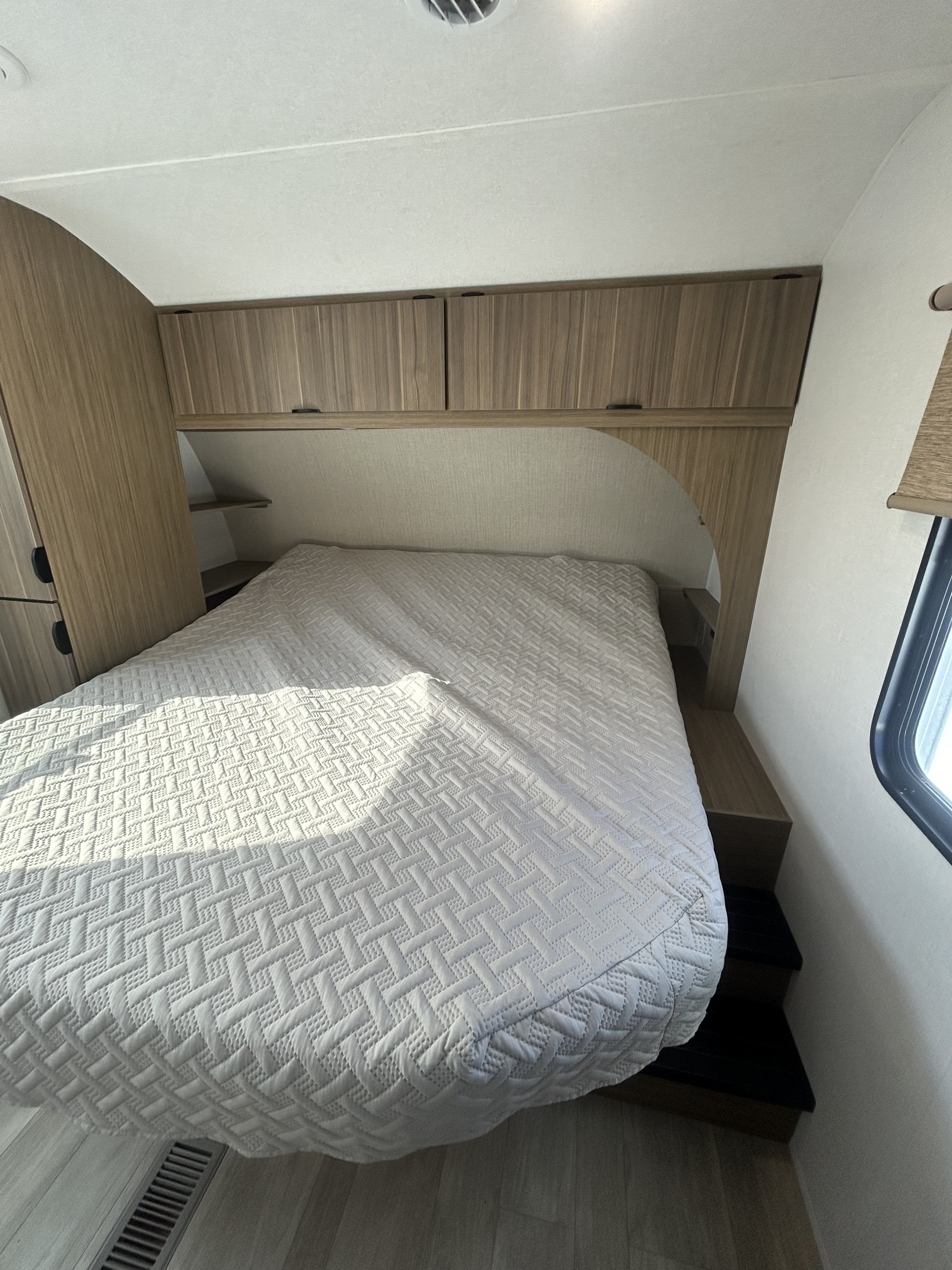 Interior view of an RV rental bedroom. A bed with a geometric patterned white comforter fills the center, flanked by wood cabinetry and shelves.. Forest River Salem 2025