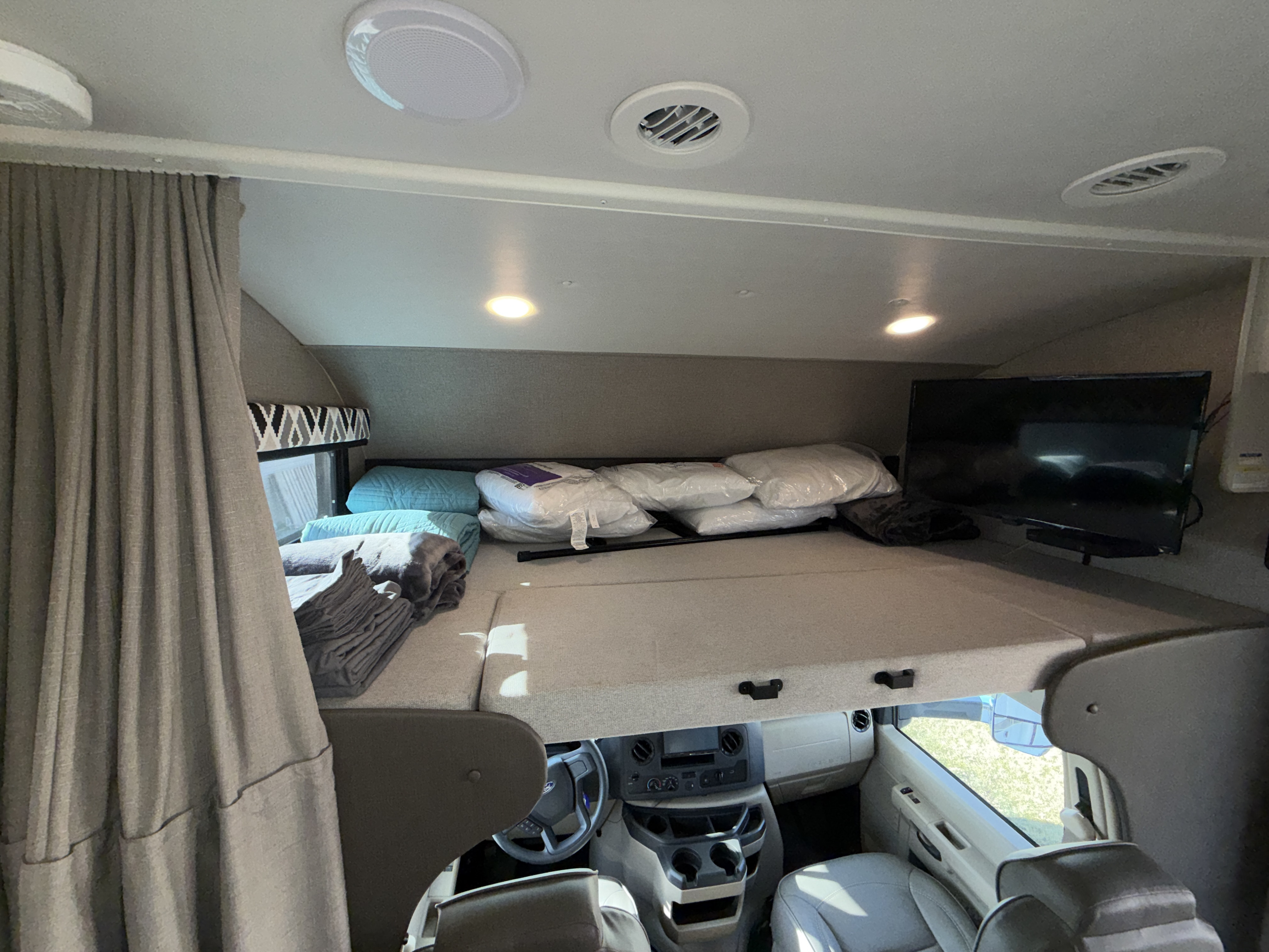 Cozy RV rental interior featuring a cab-over bunk with bedding, a TV, and the driver's cockpit visible below.. Jayco Greyhawk 2021