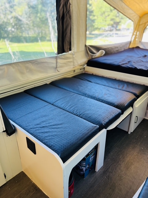 Cozy RV rental interior: Pop-up camper with dark blue cushioned benches that convert to beds, wood floor, and green outdoor view.. Coleman Grand Tour Laramie 1989