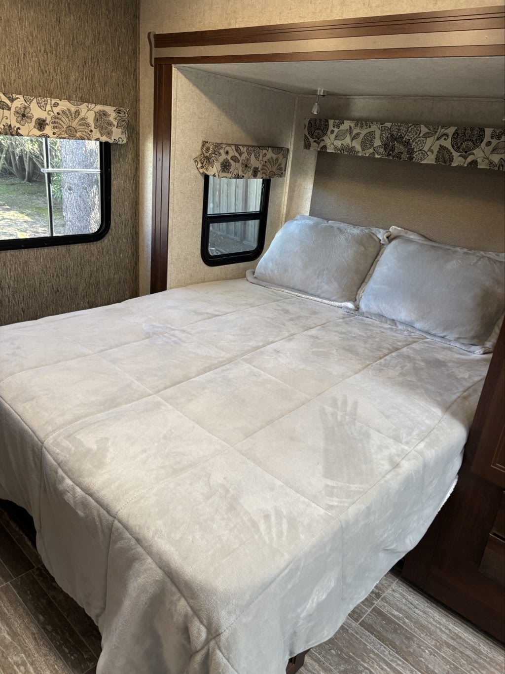 Cozy RV rental bedroom features a comfortable bed with light bedding, two small windows, and floral valances.. Forester MBS 2018