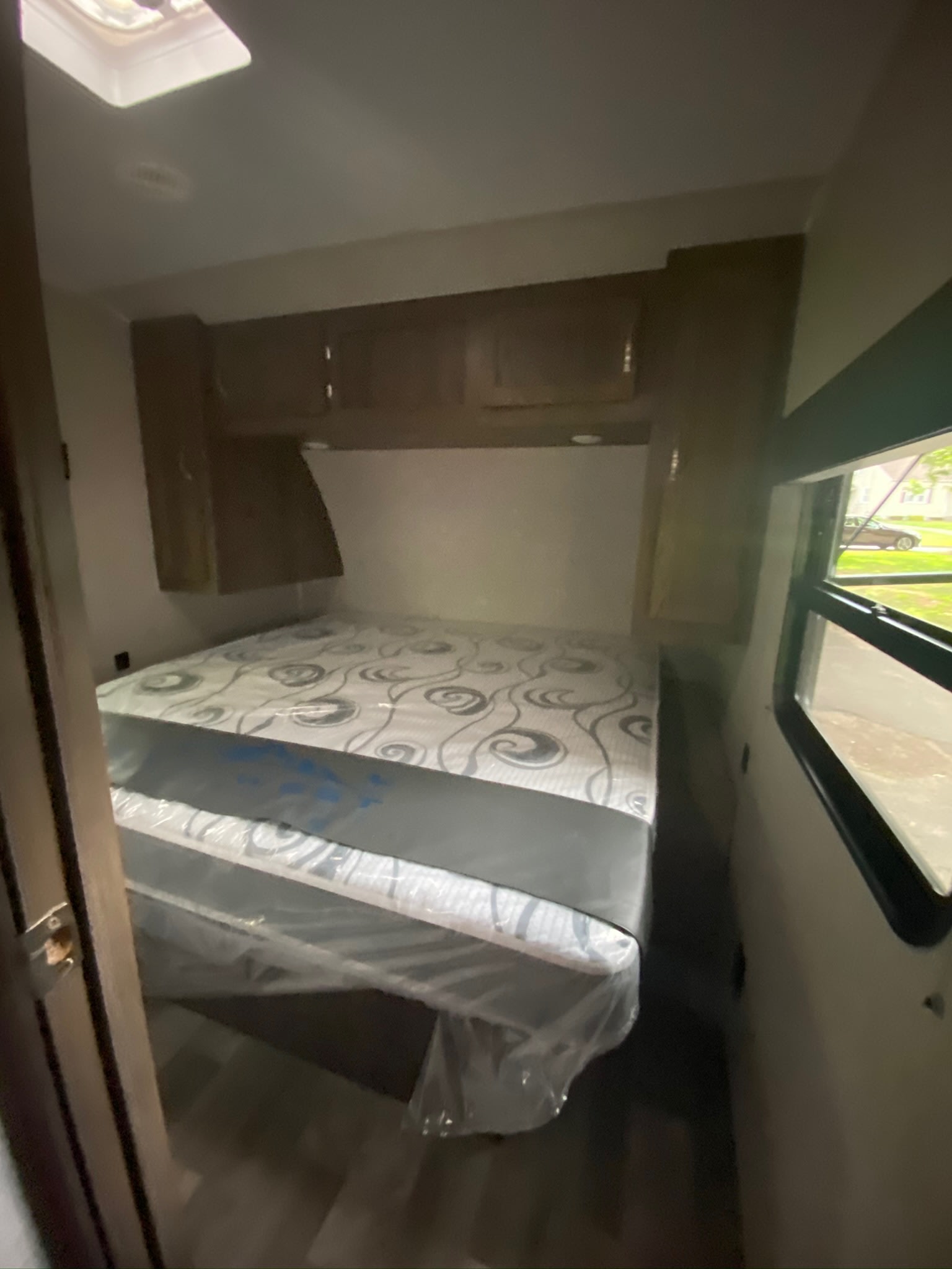 Compact rv rental bedroom with new, plastic-wrapped mattress, overhead storage, and window overlooking a street.. East to West Della Terra 2021